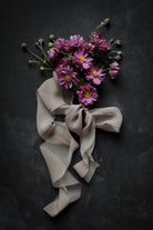 A bouquet of mauve flowers with yellow centers tied with a dusty mauve sheer silk ribbon, arranged on a dark textured background.
