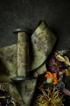 Limited-edition eco-print silk ribbon displayed on a wood spool surrounded by an assortment of dried flowers