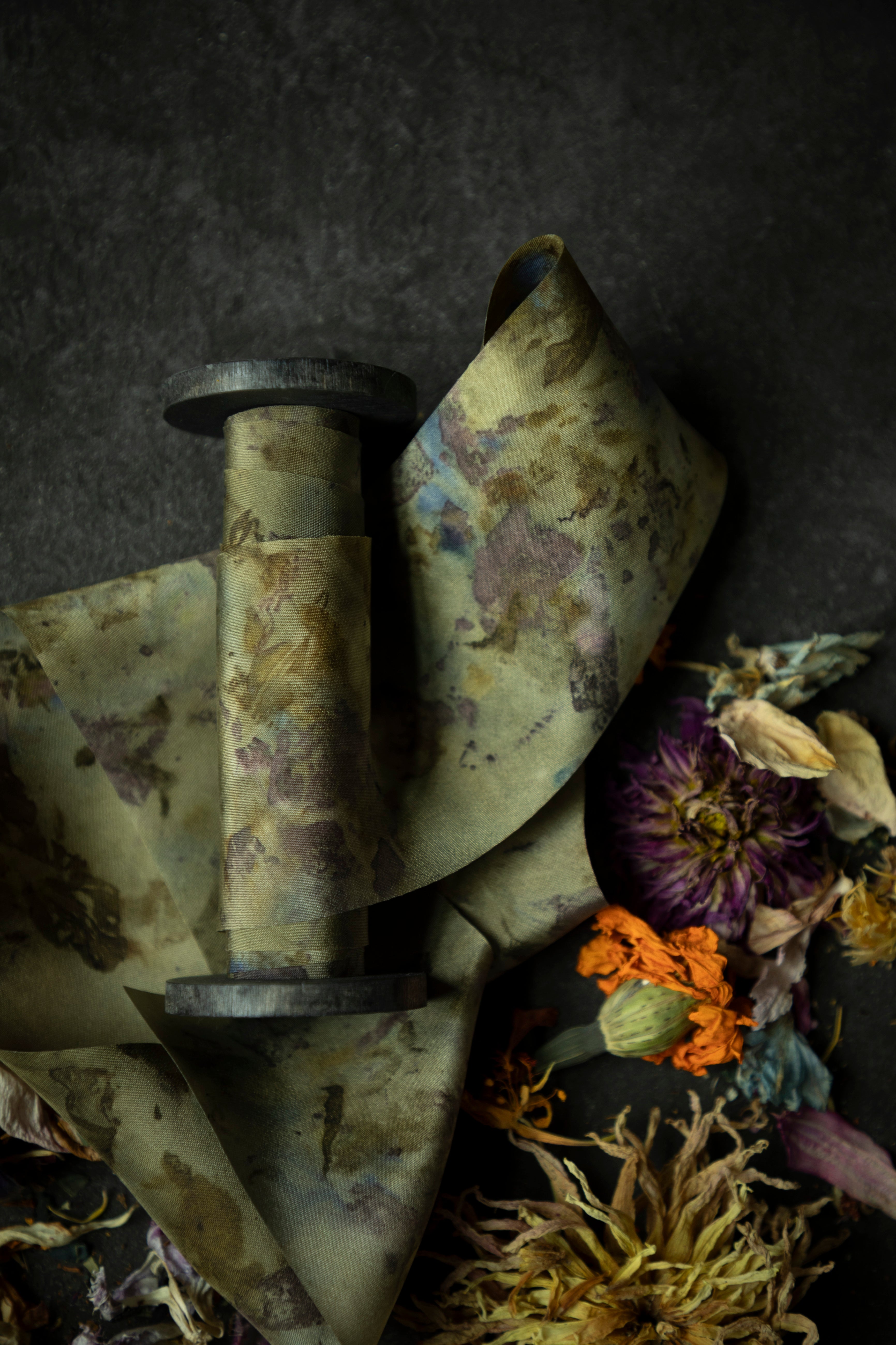 Limited-edition eco-print silk ribbon displayed on a wood spool surrounded by an assortment of dried flowers