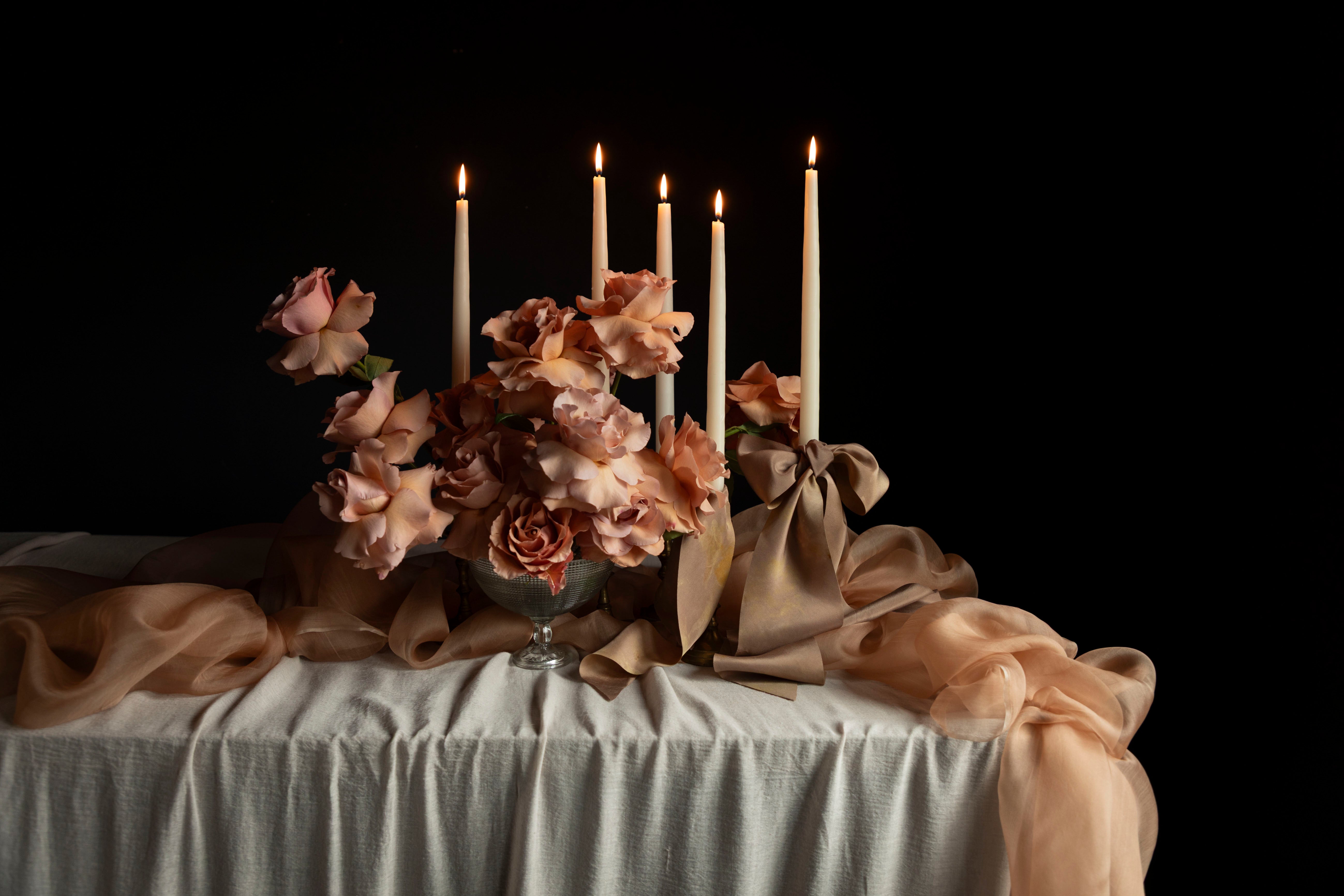 Decorative floral arrangement with coral roses and candles on a handmade tablecloth against a black background 