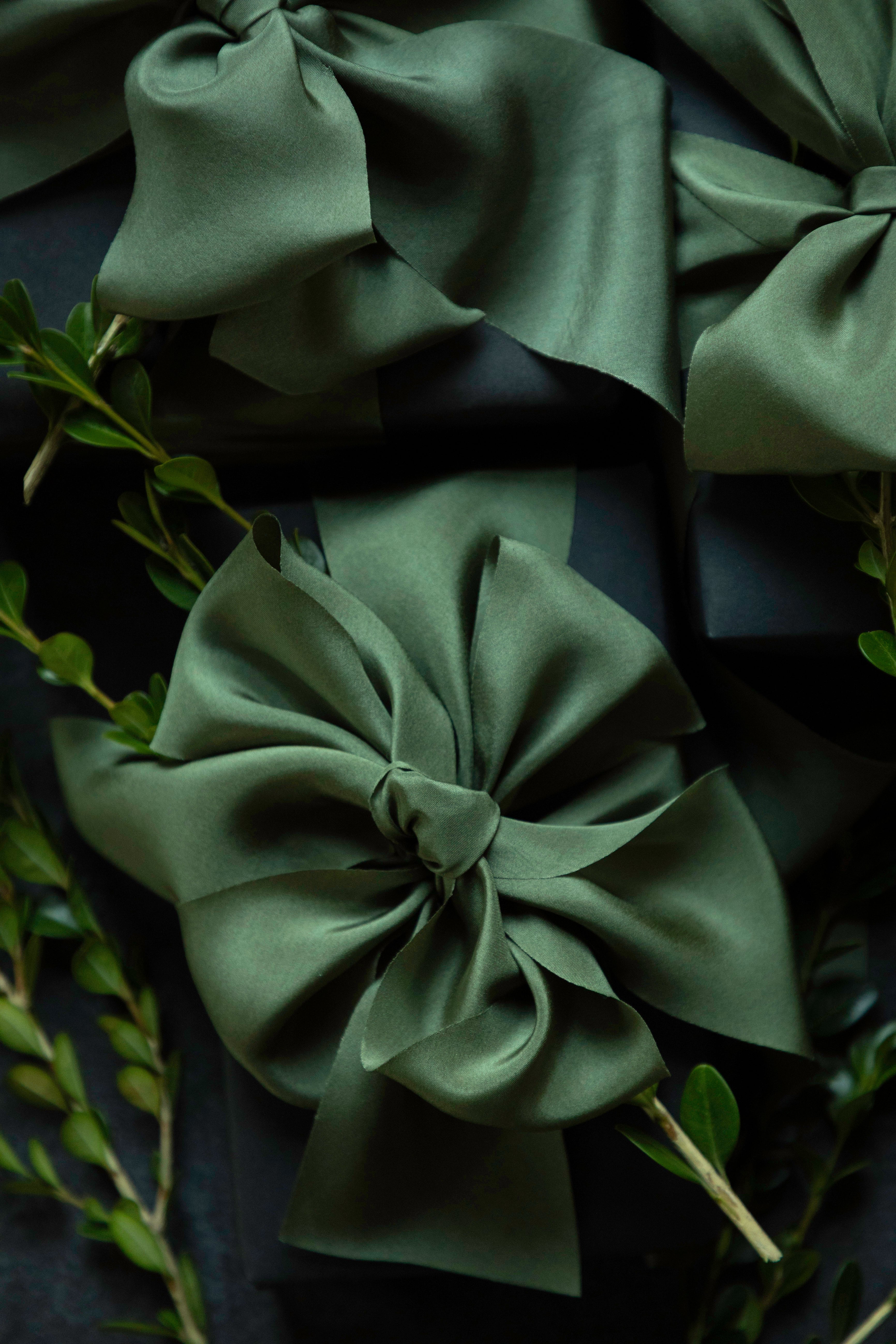 A close-up of dark olive green satin ribbon bows arranged on a dark background with small green leaves.