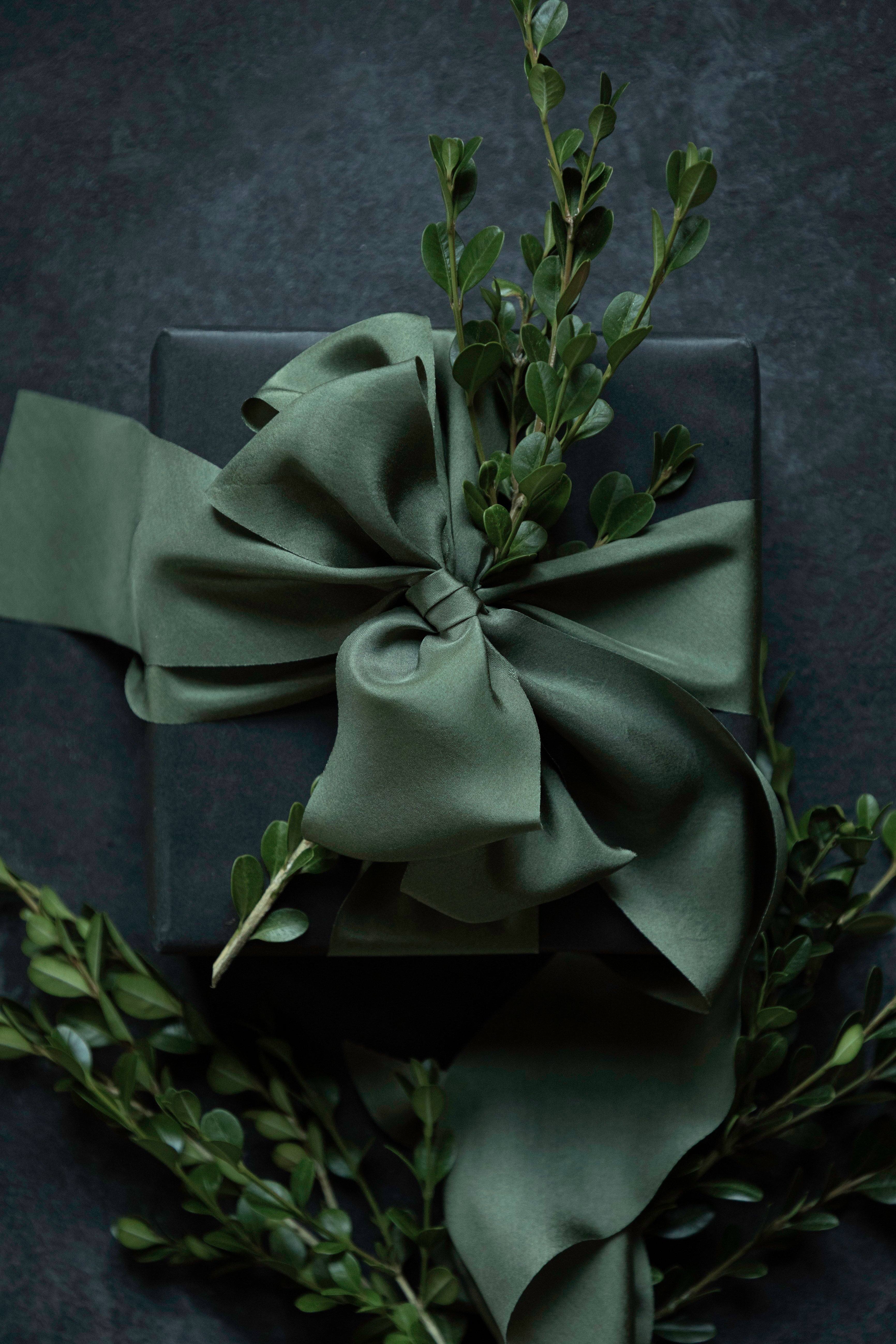 An olive green satin ribbon tied in a large bow around a dark gift box, decorated with green foliage, set against a dark textured background. Luxury gift Wrapping 