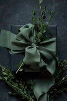 An olive green satin ribbon tied in a large bow around a dark gift box, decorated with green foliage, set against a dark textured background. Luxury gift Wrapping 
