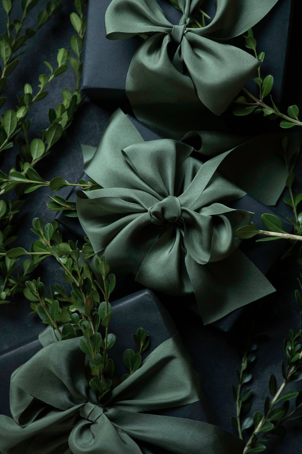 Three dark green satin gift bows tied around dark navy wrapped boxes, styled with green foliage on a dark background.