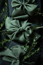 Three large satin gift bows in a muted sage green color, tied around dark wrapped boxes and accented with delicate green foliage. Gift wrapping inspiration