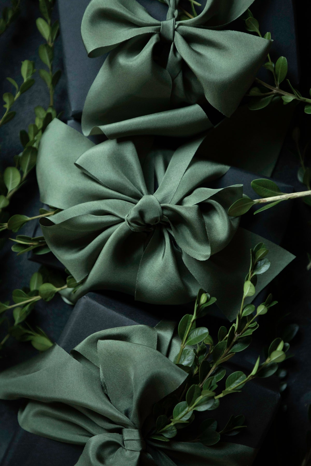 Three large satin gift bows in a muted sage green color, tied around dark wrapped boxes and accented with delicate green foliage. Gift wrapping inspiration