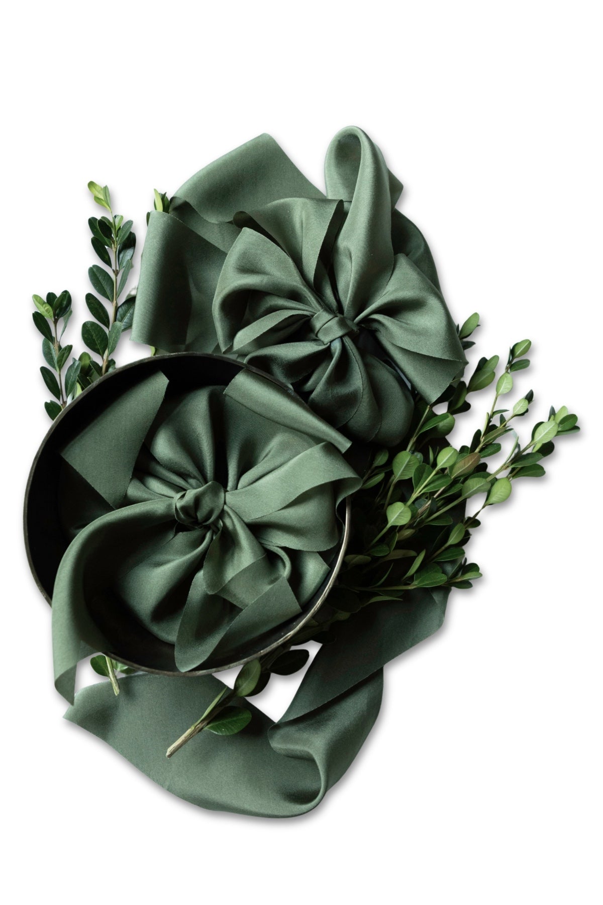 A dark sage green satin ribbon arranged in two large bows, styled with green boxwood branches on a plain white background.