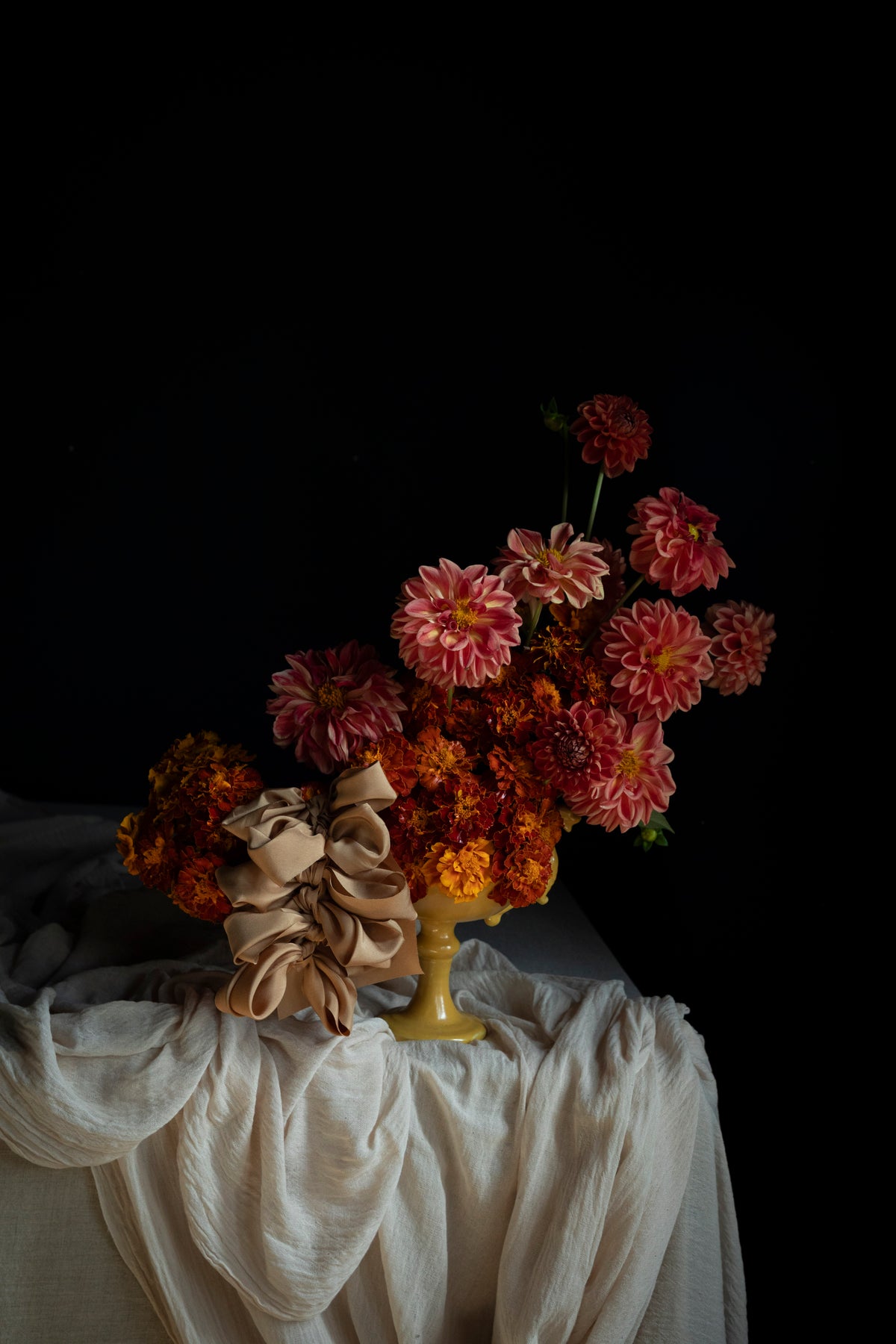 A still life floral arrangement featuring pink and orange dahlias with a beige ribbon bow, displayed in a gold pedestal vase draped with white fabric against a solid black background.