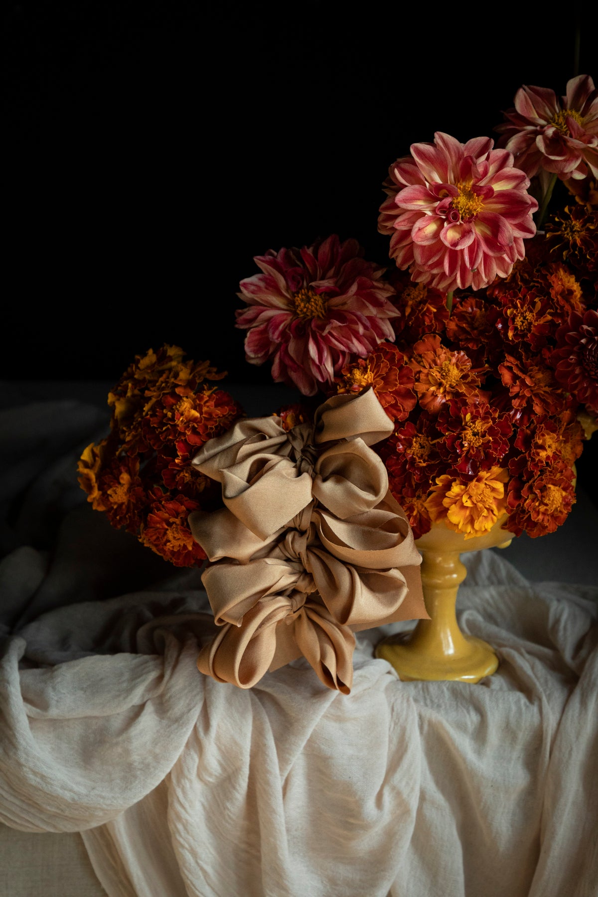 A styled still life featuring a large cinnamon-coral silk ribbon bow in the foreground, with a bouquet of orange, dahlia and marigold flowers in a yellow vase. On a draped white fabric against a black background.