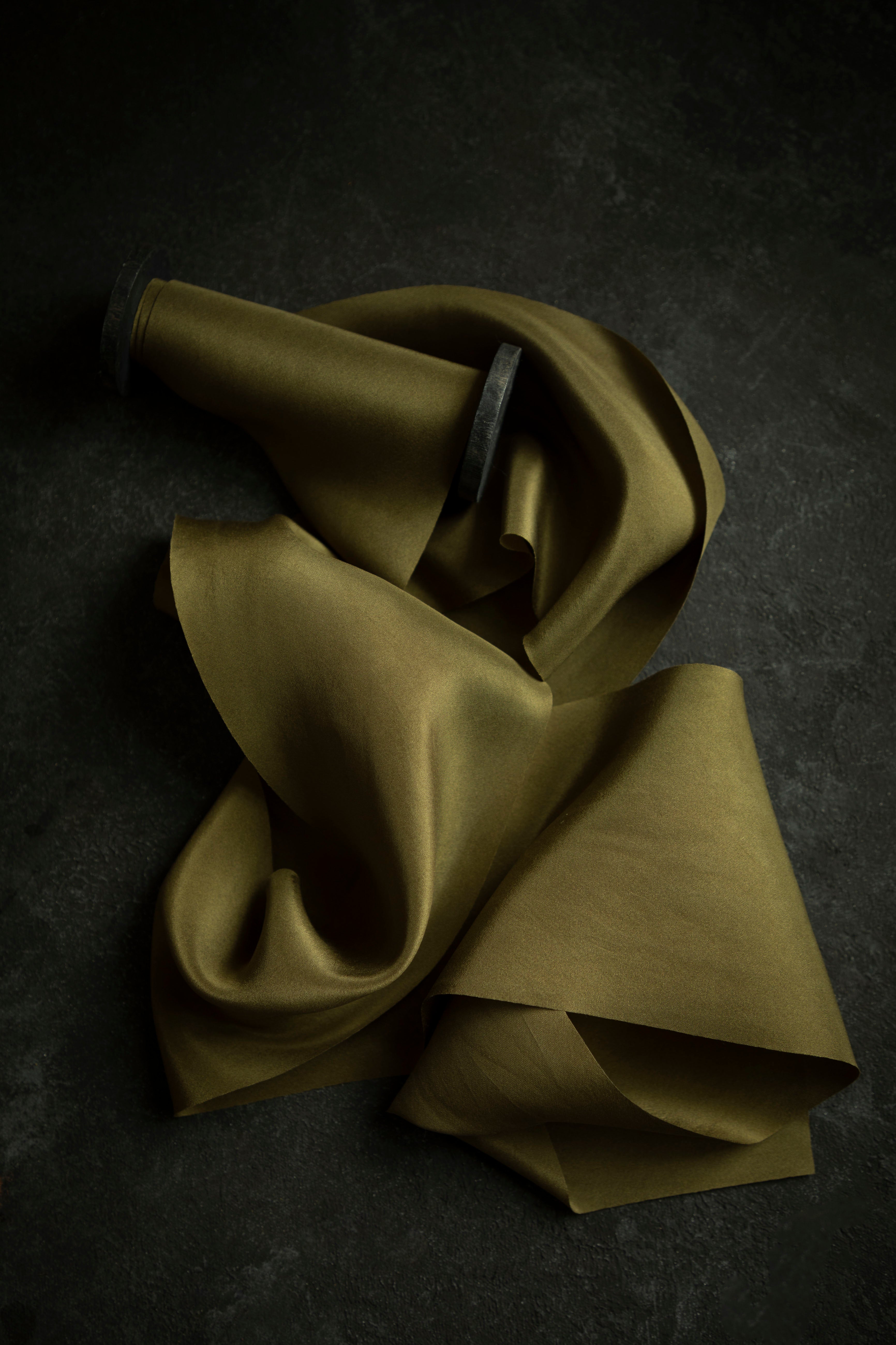 A Forest green silk ribbon with subtle olive-yellow undertones, laying on a dark surface, demonstrating its elegant and sophisticated texture.