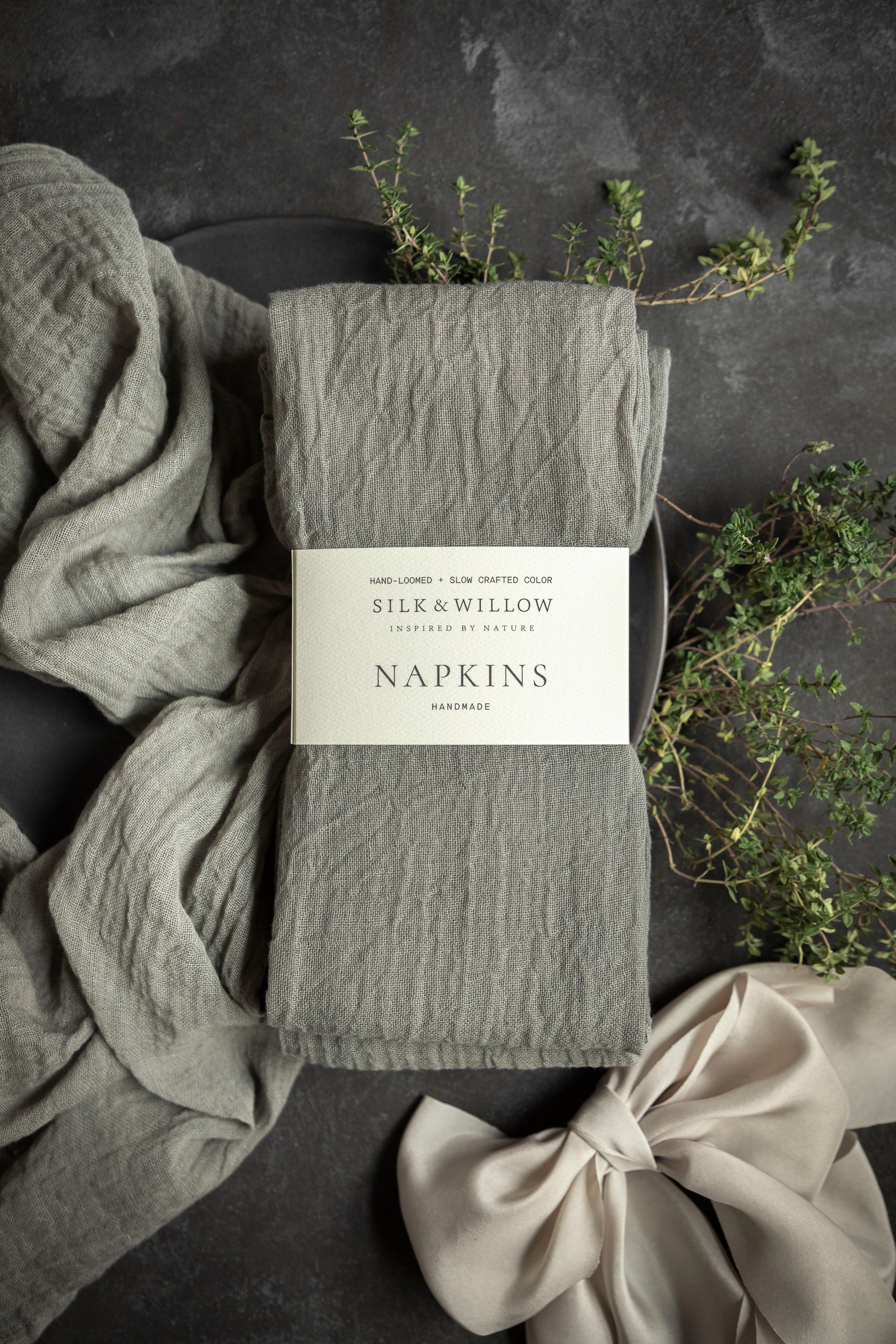 Pack of gray napkins with a brand label on a dark surface with fabric and plants.