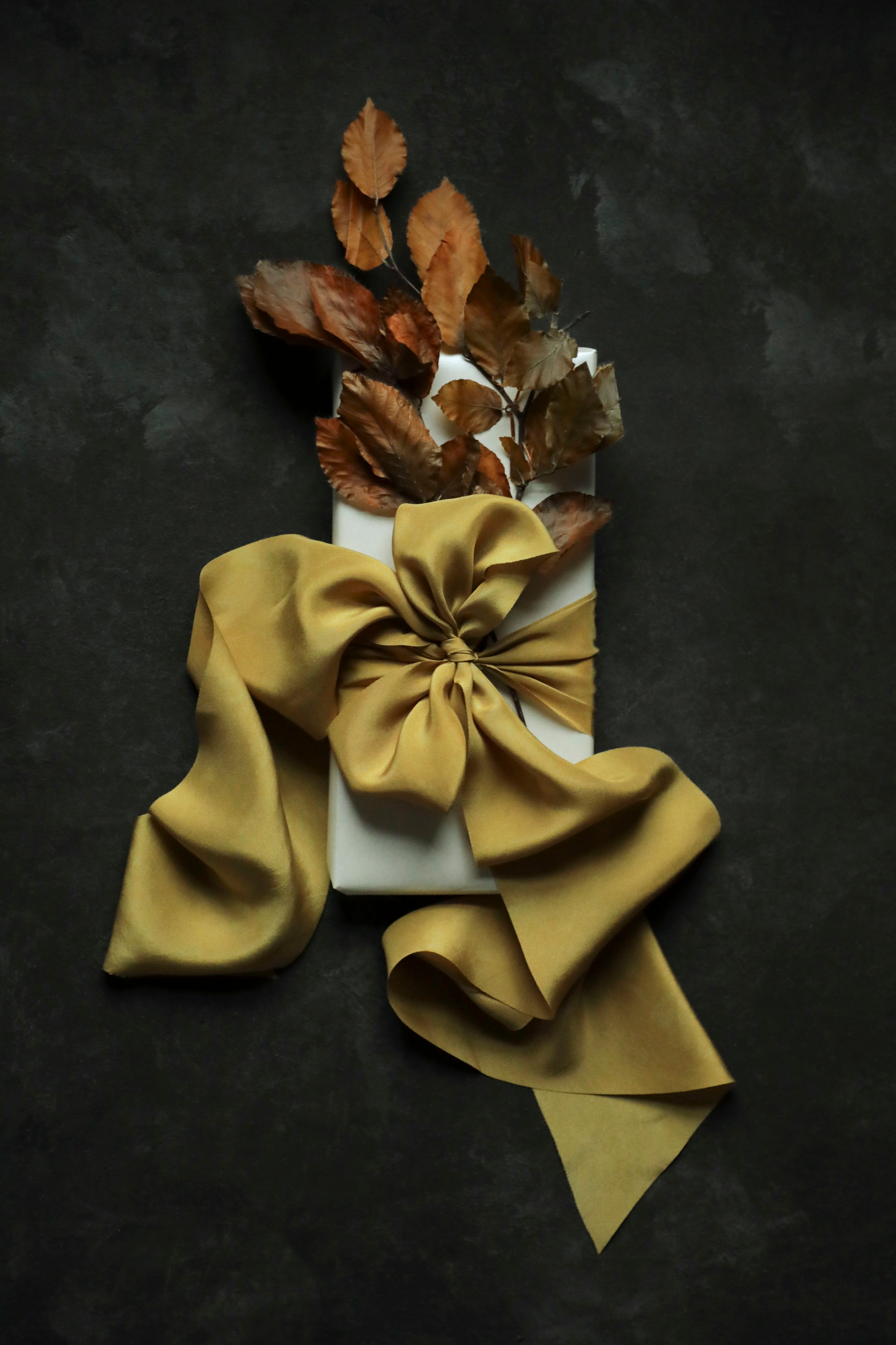 A white rectangular box wrapped with a large satin gold ribbon and bow, topped with dried brown leaves, arranged on a dark textured background.