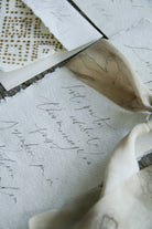 A close-up of layered cream and beige fabrics with delicate cursive script and a small geometric dot pattern, arranged on a rustic wooden surface.