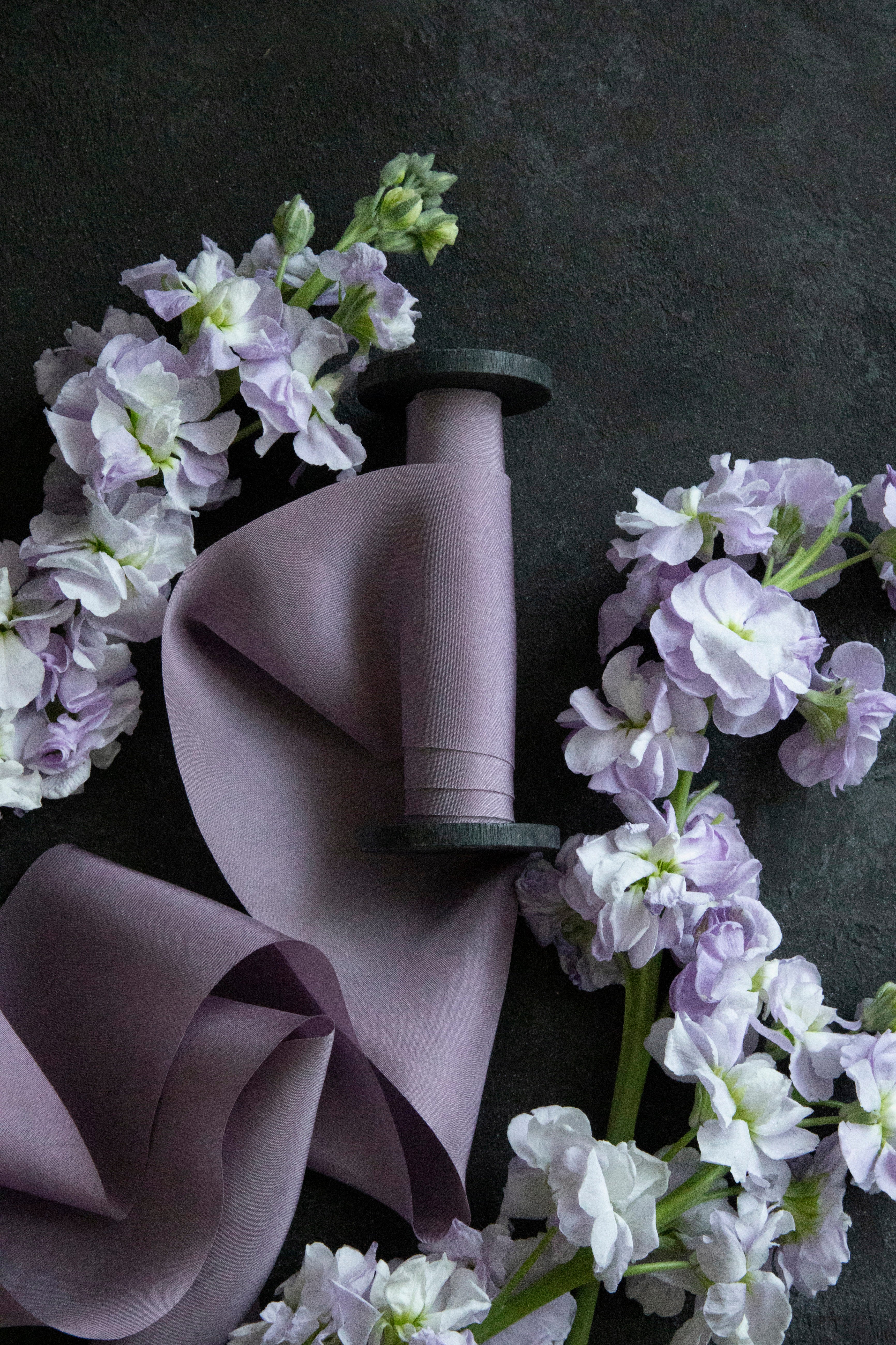 A medium-tone purple lilac silk ribbon is displayed alongside some lilac flowers on a dark textured background, with a spool of ribbon partially visible.