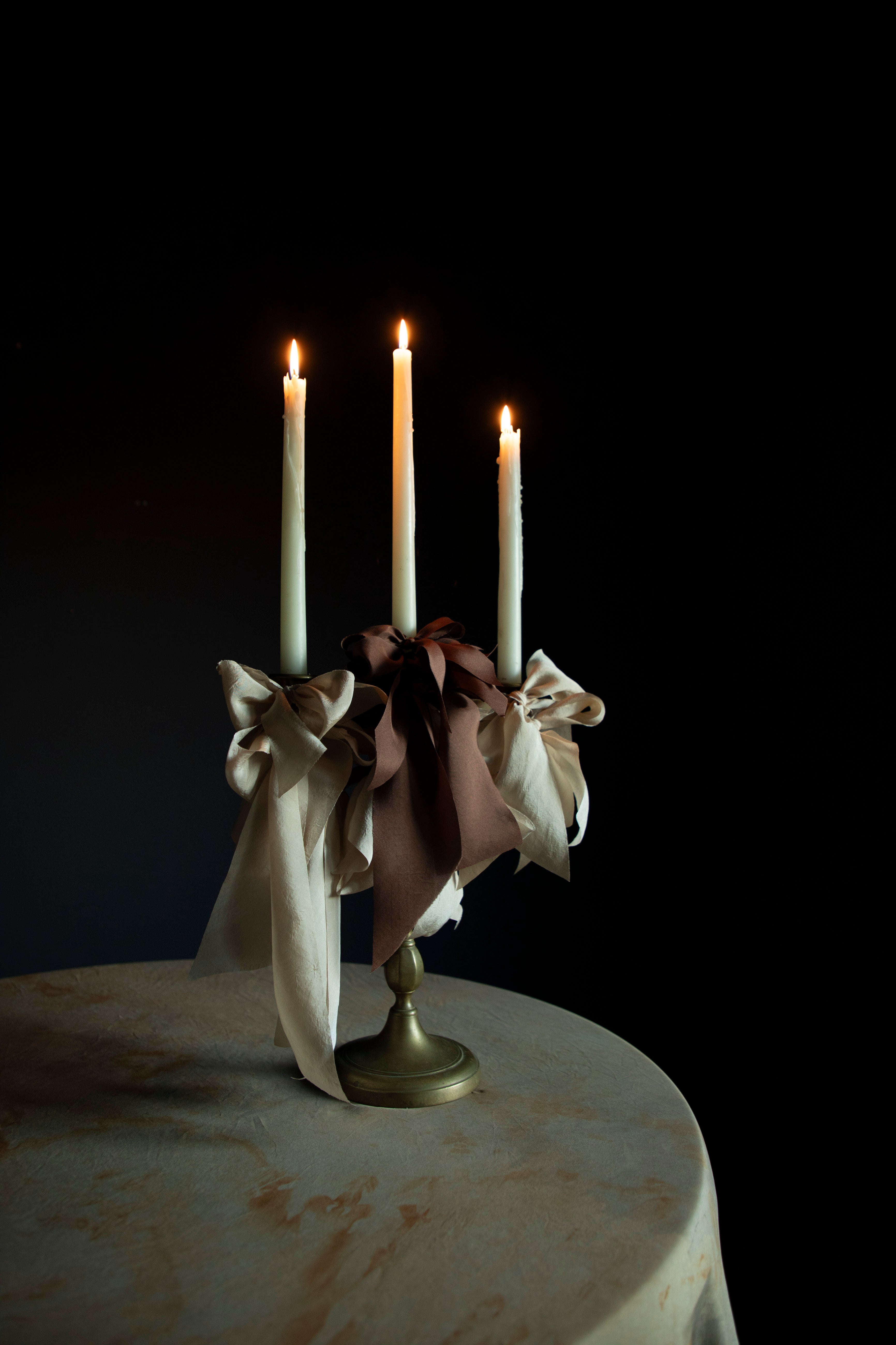 A mocha silk ribbon tied in a bow around a candlestick holder with lit candles, set against a dark background.