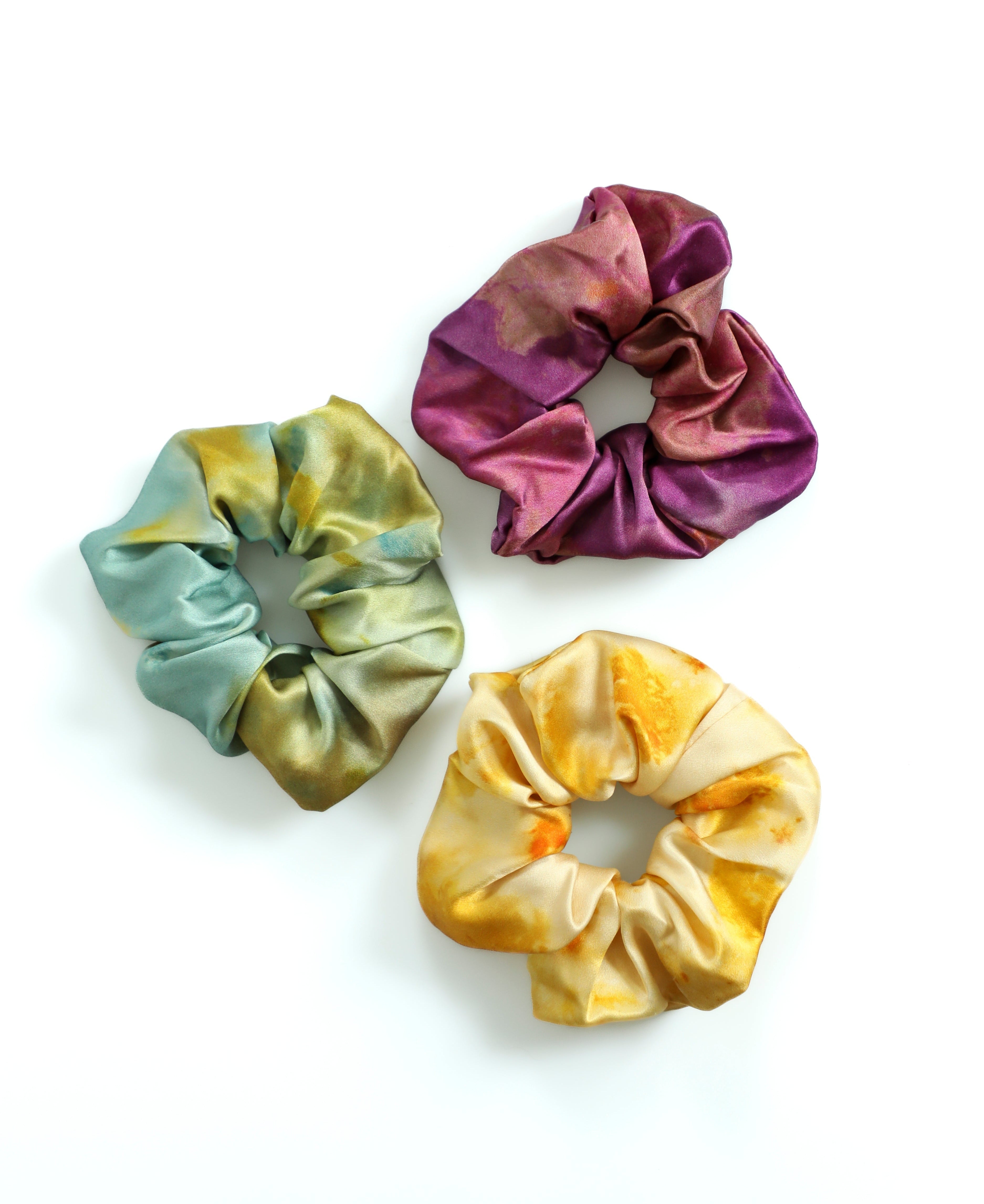 Three tie-dye scrunchies in purple, green, and yellow on a white background
