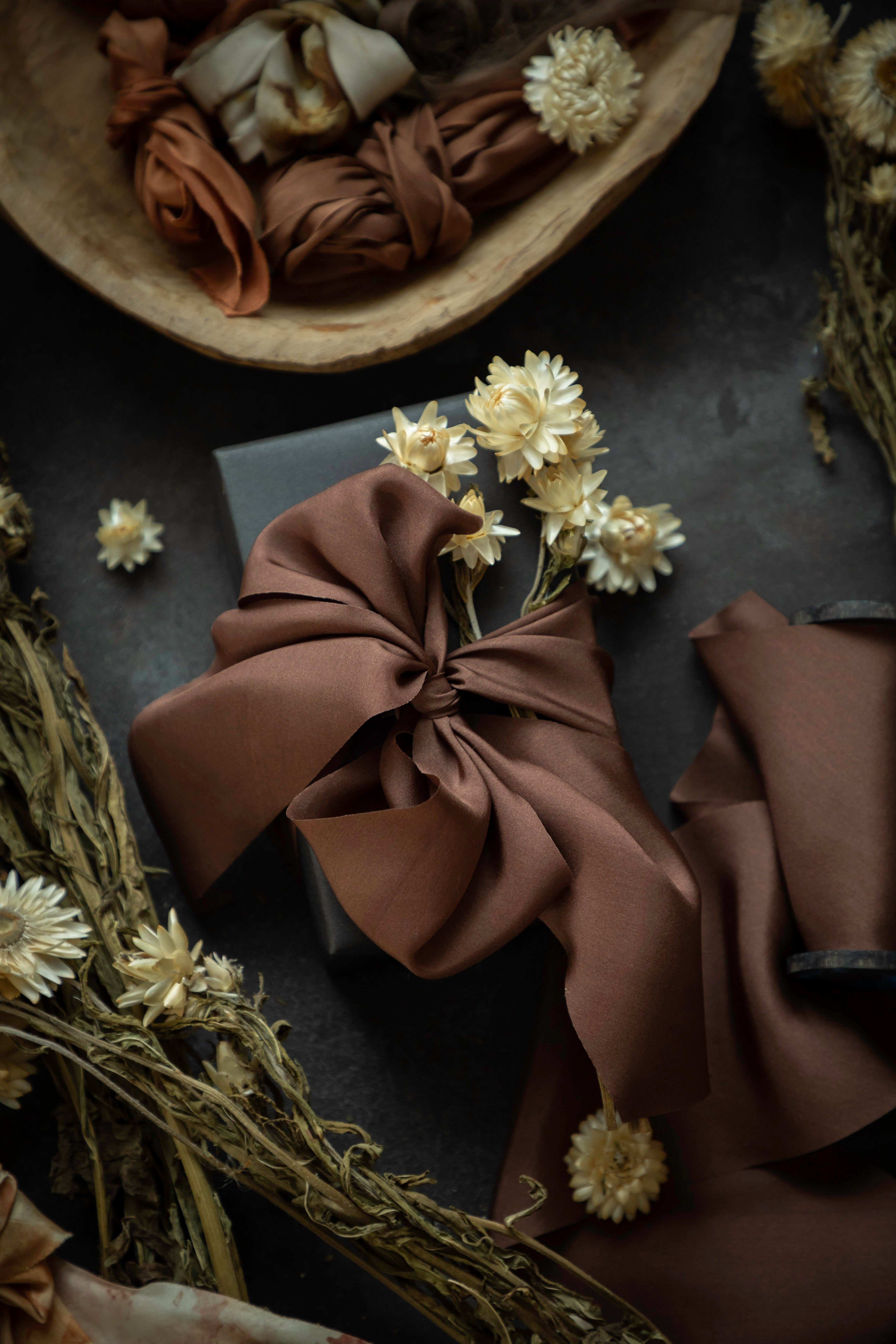 A styled flat-lay of a brown silk ribbon tied in a large bow around a dark box, surrounded by dried flowers and neutral-toned fabric pieces on a dark textured surface.
