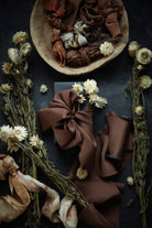 A styled flat-lay of brown silk ribbon or fabric in multiple tied bows, arranged with dried white flowers on a dark textured surface. A wooden bowl at the top contains additional folded brown fabric, and more ribbon and floral props extend across the frame.
