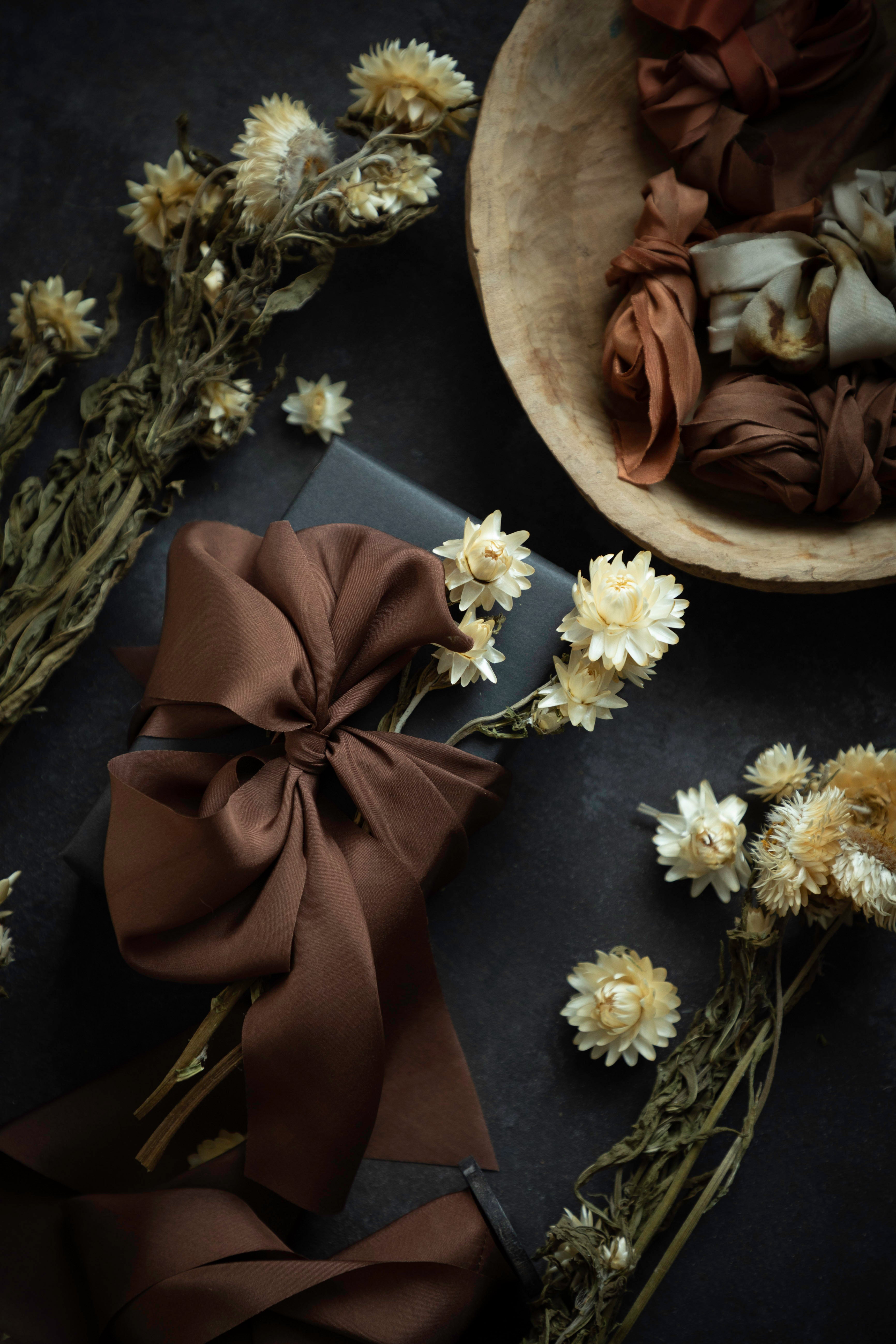 A styled flat-lay featuring a large, glossy dark brown satin ribbon bow on a dark surface, surrounded by dried cream flowers and a wooden bowl filled with more brown and beige ribbons.