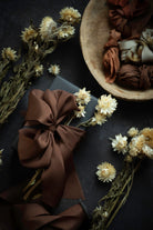 A styled flat-lay featuring a large, glossy dark brown satin ribbon bow on a dark surface, surrounded by dried cream flowers and a wooden bowl filled with more brown and beige ribbons.