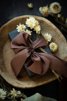 A dark gray gift box wrapped in a large brown satin bow and decorated with dried cream flowers, presented on a rustic wooden plate against a dark background.