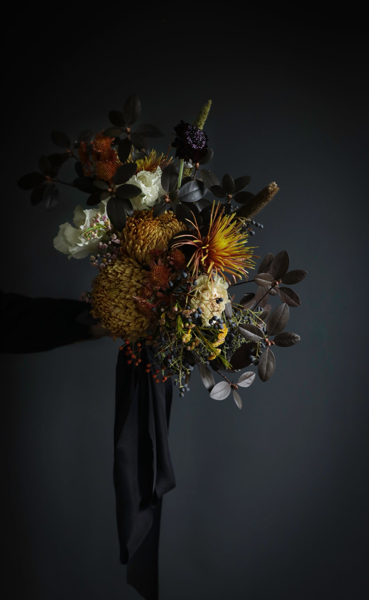 A rustic flower bouquet featuring a mix of orange, yellow, white, and dark brown botanicals with a long black ribbon, photographed against a dark gray background.
