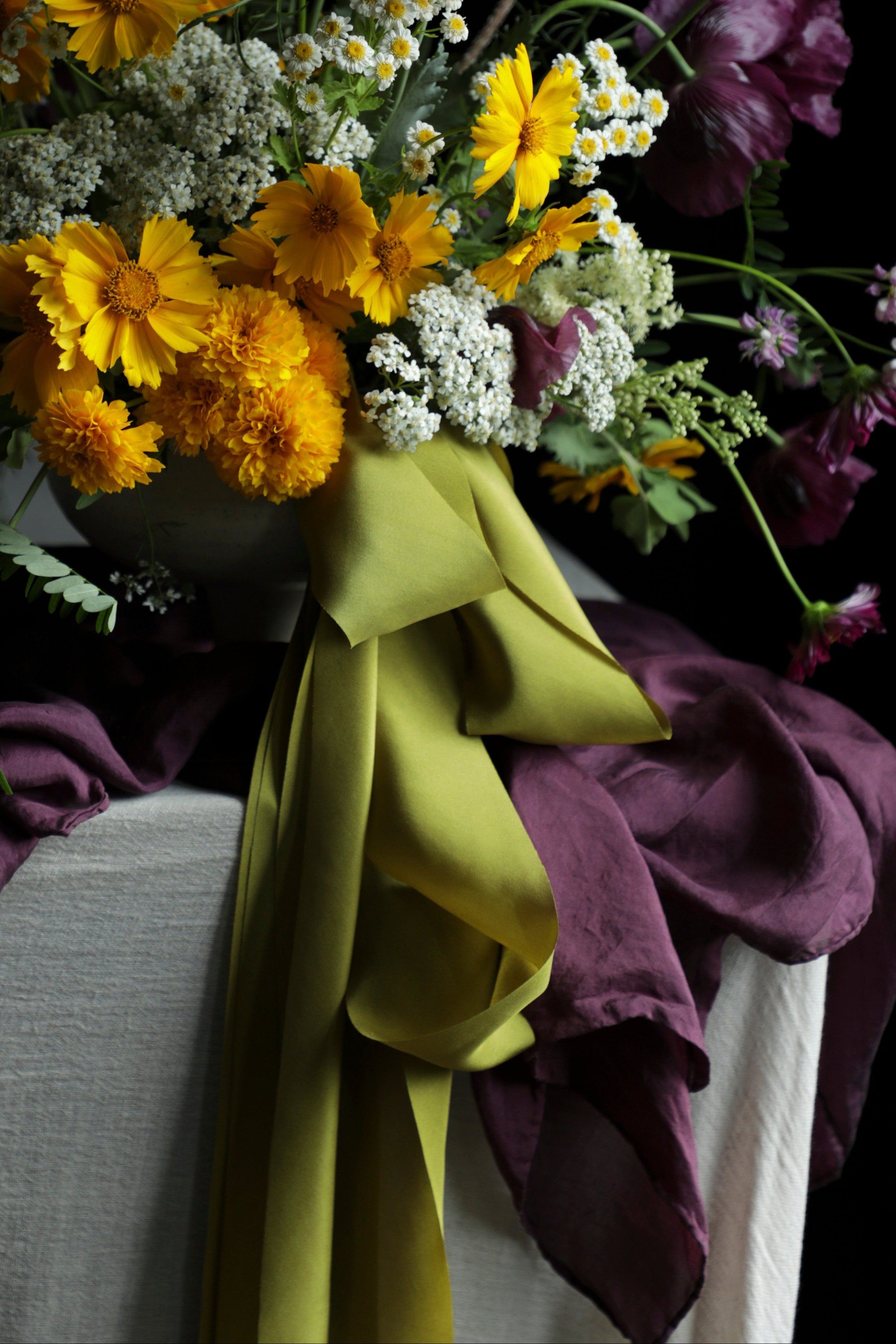 A rustic wildflower bouquet in a dark vase, featuring yellow daisies, purple poppies, white baby's breath, and a large olive green ribbon bow, set against a dark background with draped purple fabric.