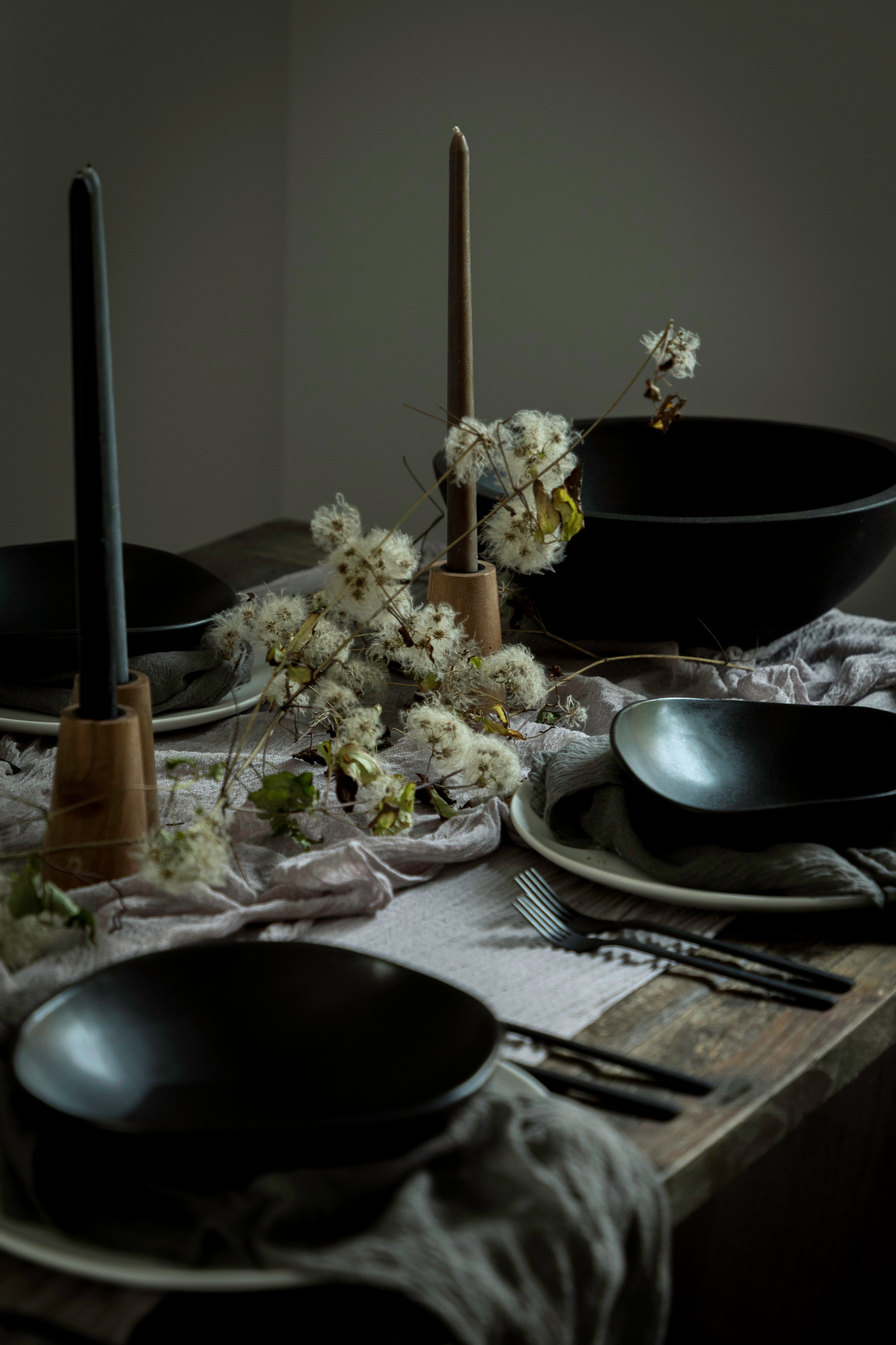 A styled dining table setting with matte black bowls, plates, black taper candles in wooden holders, and dried white flowers on a rustic wooden table with draped linen.