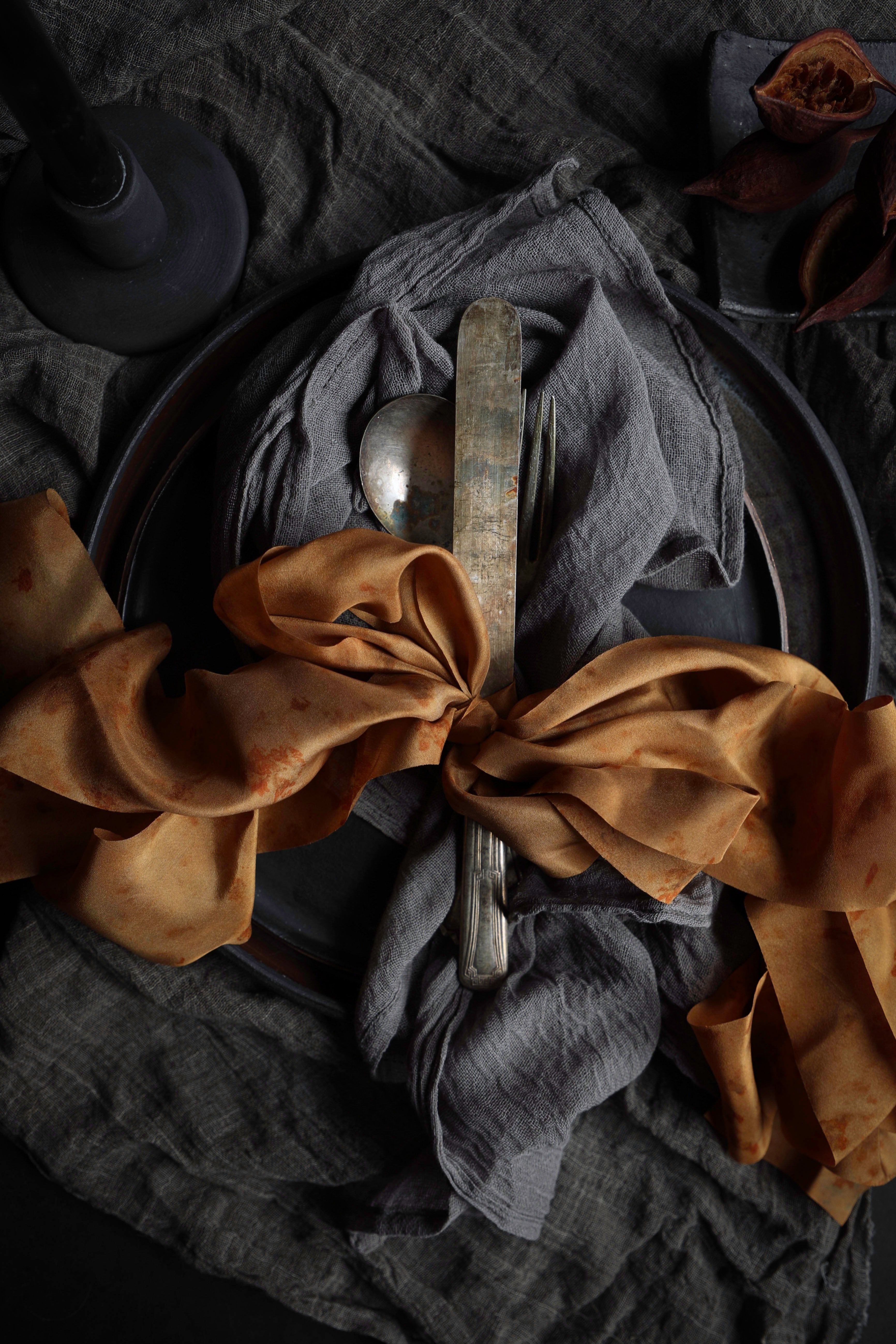 A styled tabletop setting with a dark gray linen napkin tied in a large bow with rust-brown fabric, a silver knife, and a spoon on a dark plate over a textured dark gray cloth.