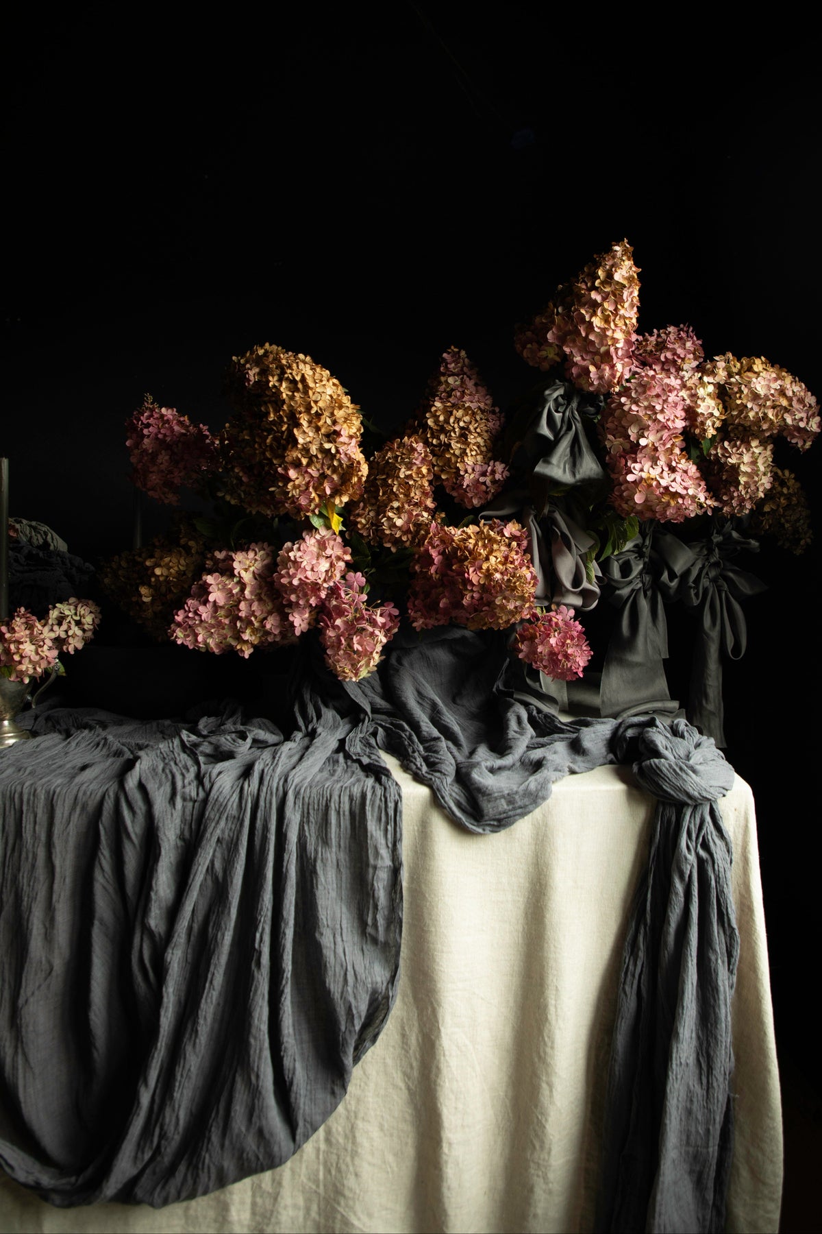 A still life arrangement of dried pink and brown flowers draped over a table covered with dark gray and off-white fabric, set against a solid black background.