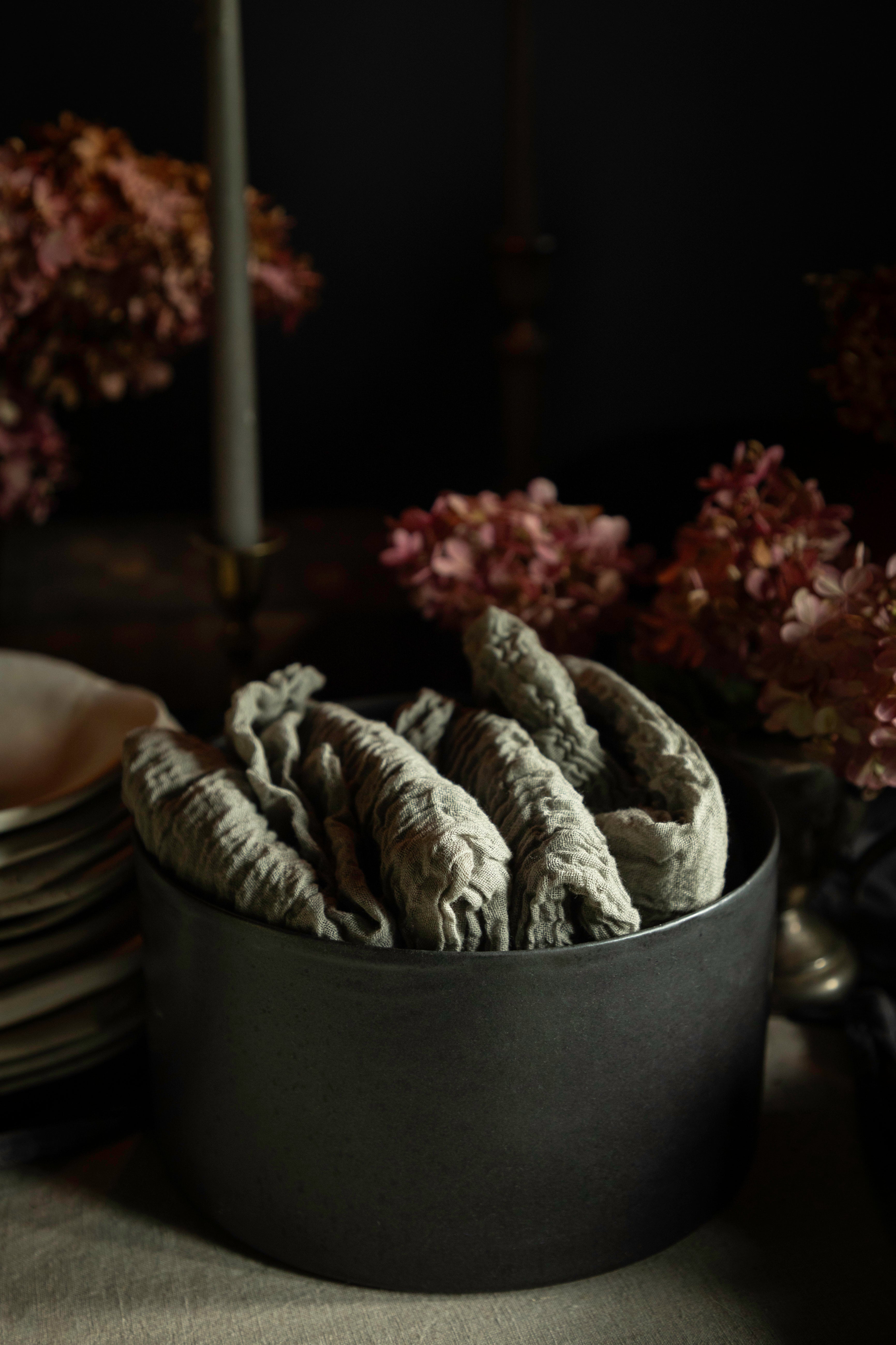 A dark round container filled with dried, wrinkled sage leaves, styled on a table with plates and pink hydrangeas in a dim, moody setting.