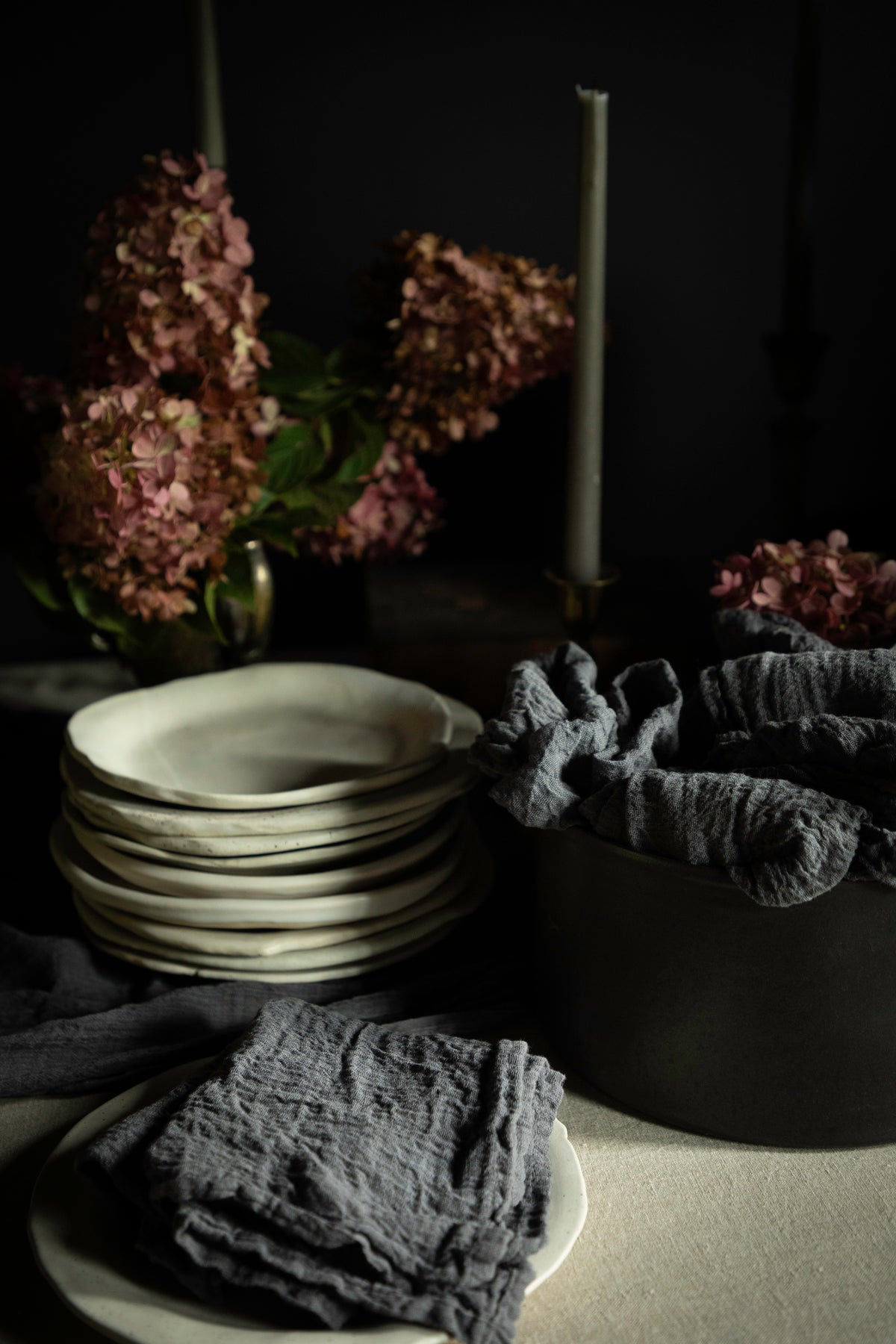 A styled tabletop scene with a stack of matte off-white ceramic plates, dark gray textured cloth napkins draped over a black bowl, and dried pink hydrangea flowers in the background against a dark backdrop.