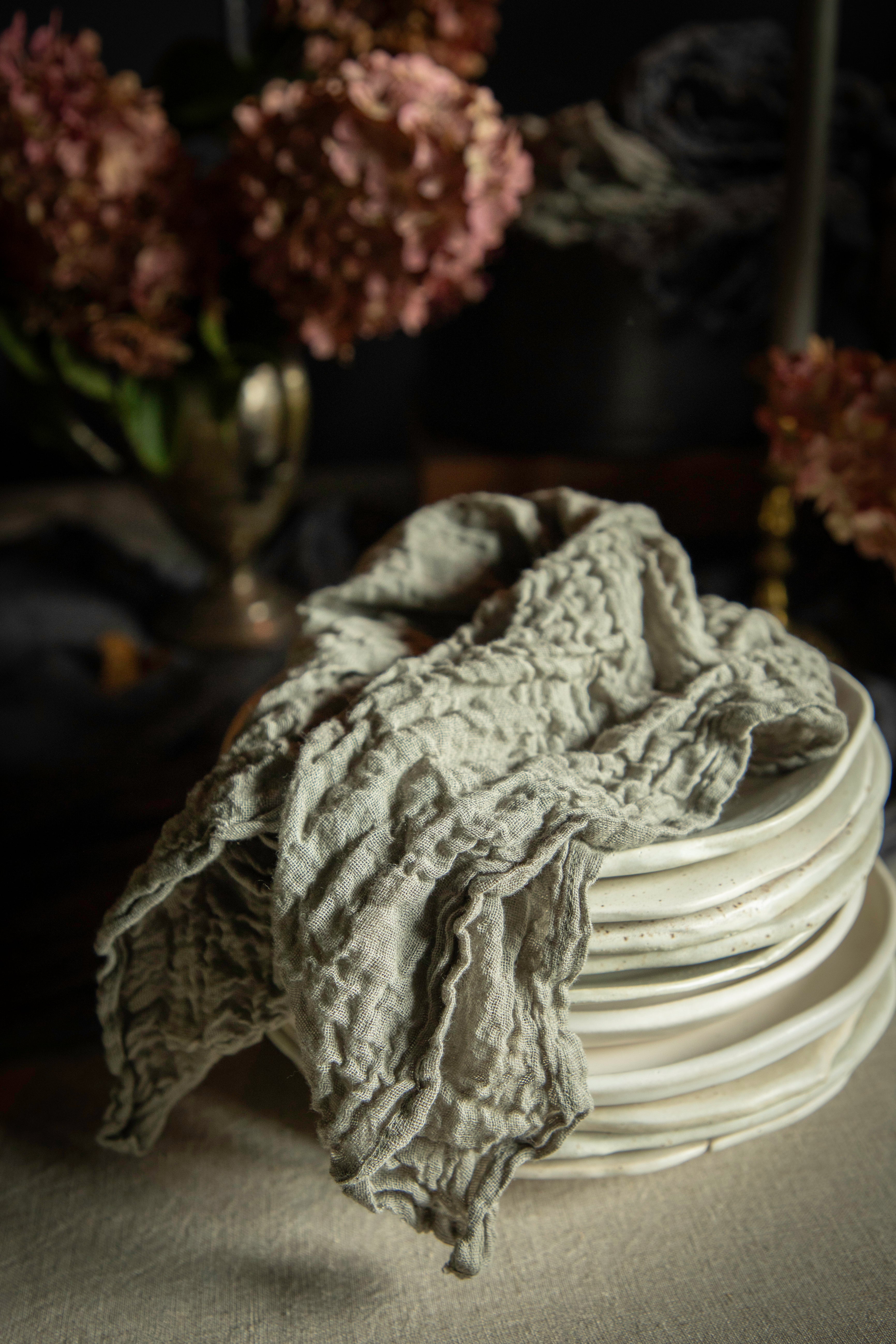 A textured, crinkled light gray linen cloth draped over a stack of white ceramic plates, set against a dark, moody background with blurred pink flowers.