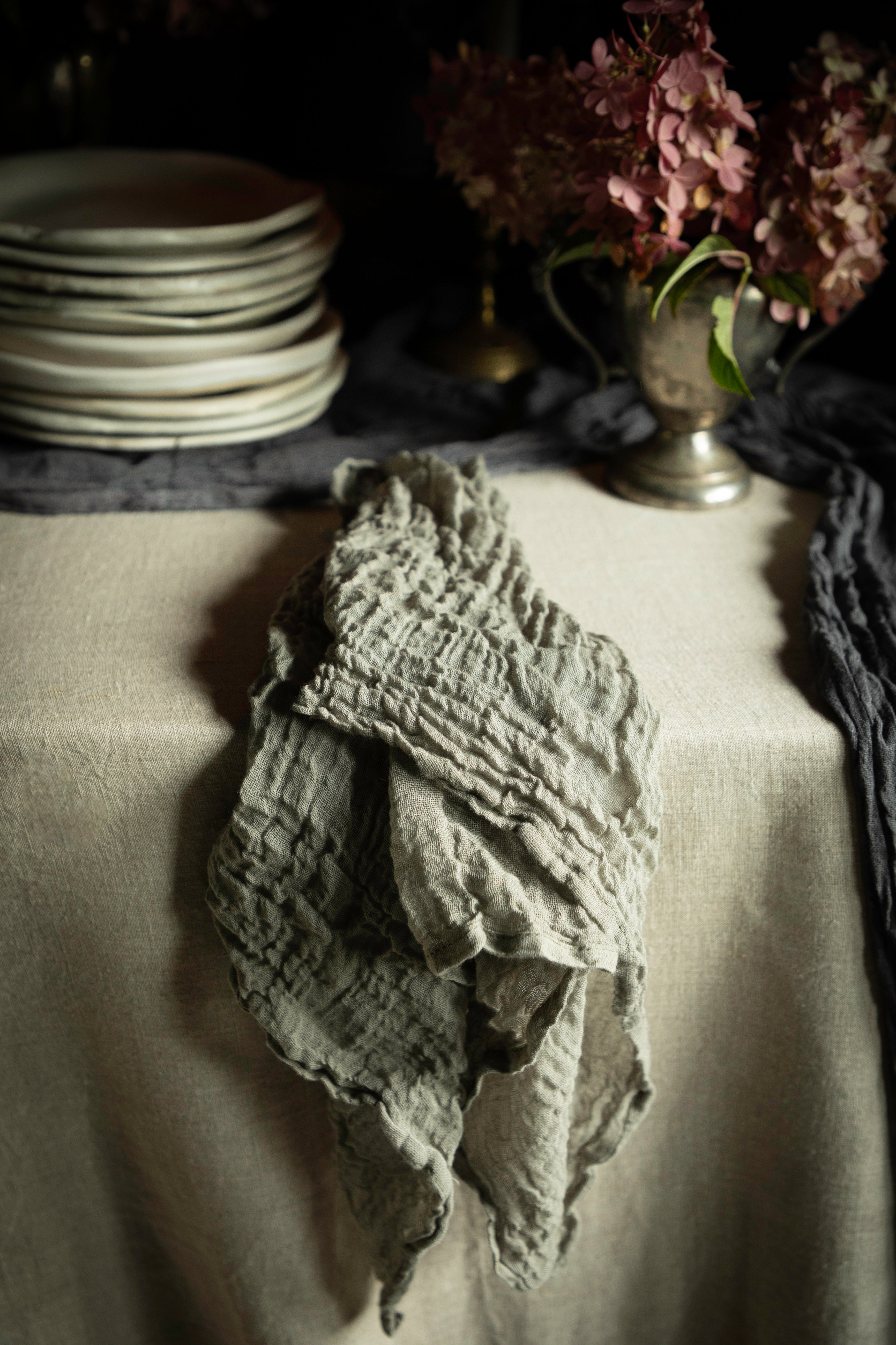 A crinkled, textured gray linen napkin draped over a beige tablecloth in a moody, rustic setting with plates and flowers in the background.