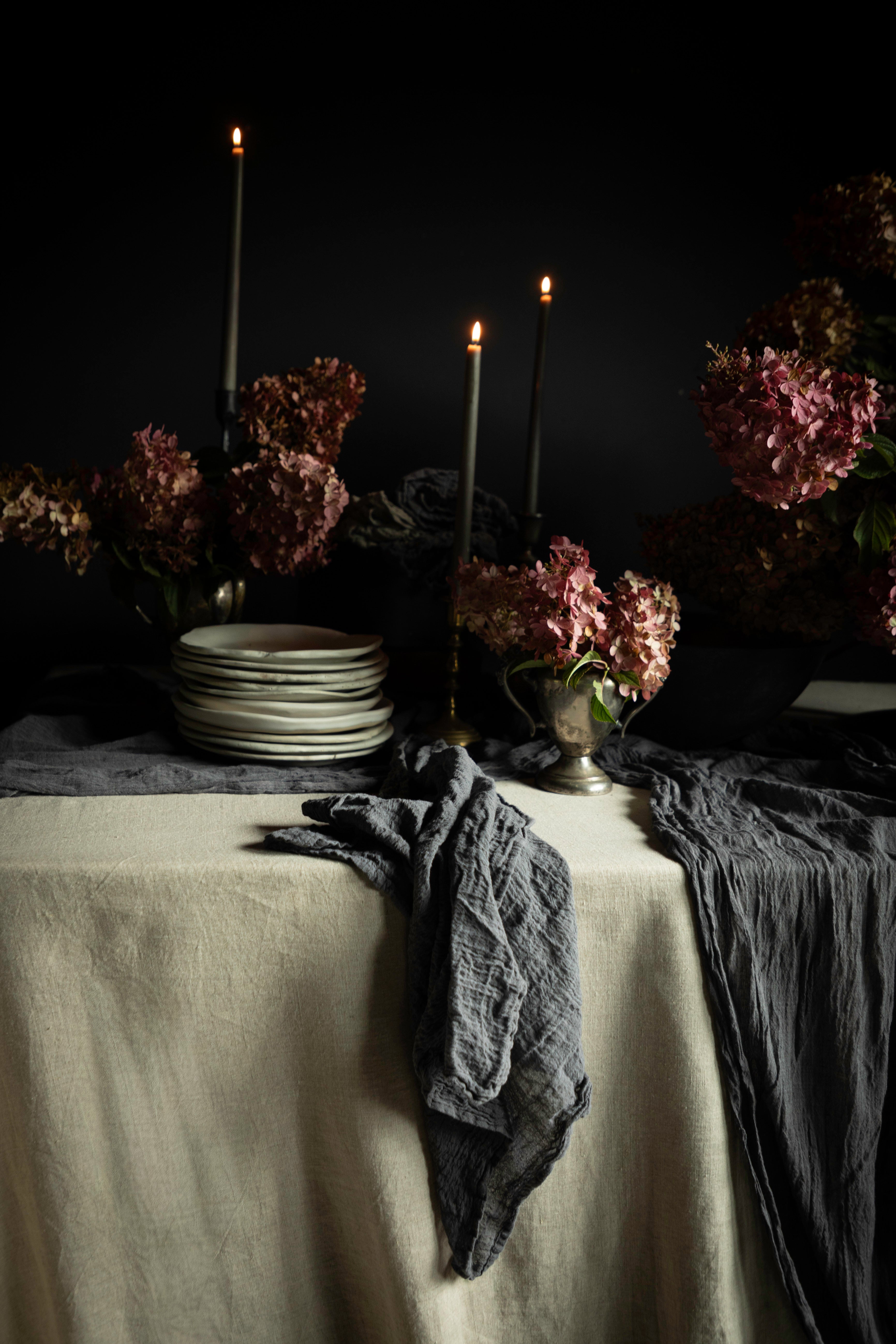 A moody tabletop still life with a cream tablecloth, dark gray crinkled napkins, stacked white plates, tall black candlesticks with lit candles, and dried pinkish-brown flowers against a black background.