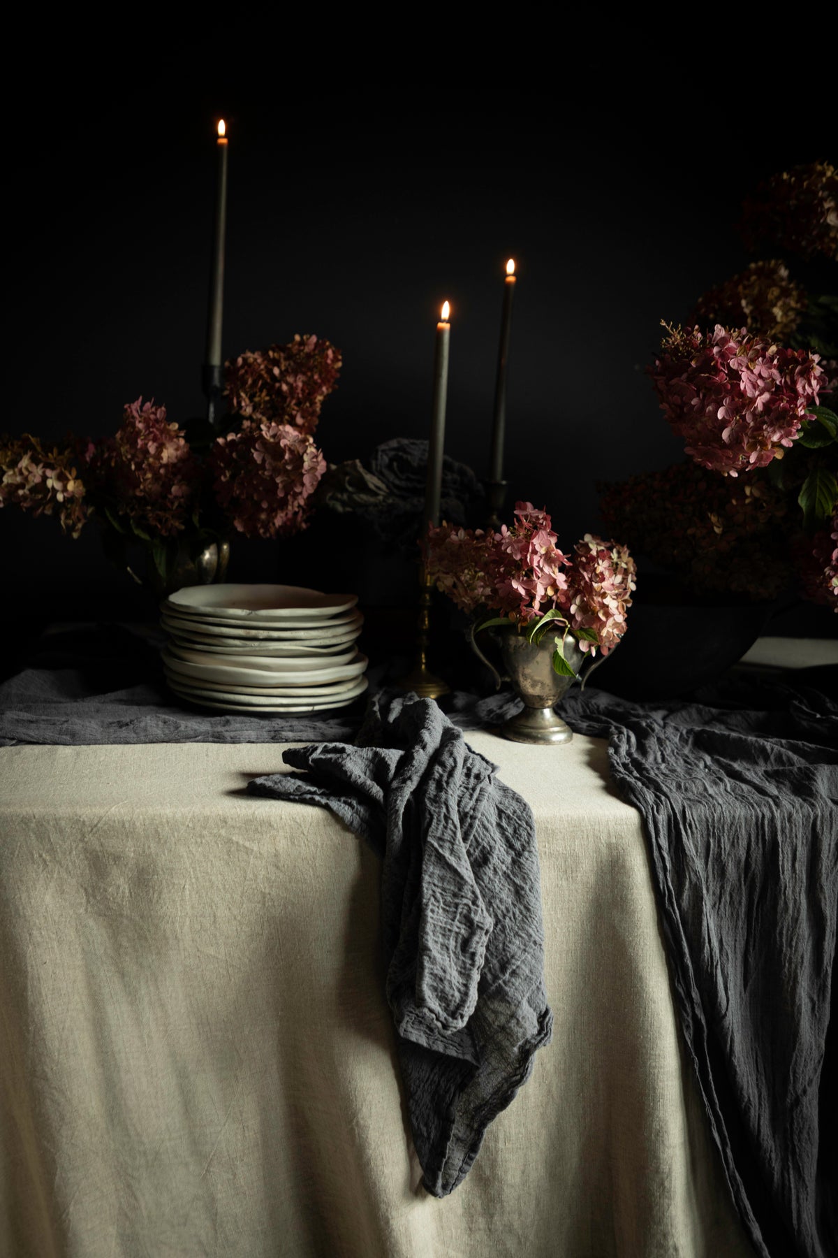 A moody tabletop still life with a cream tablecloth, dark gray crinkled napkins, stacked white plates, tall black candlesticks with lit candles, and dried pinkish-brown flowers against a black background.
