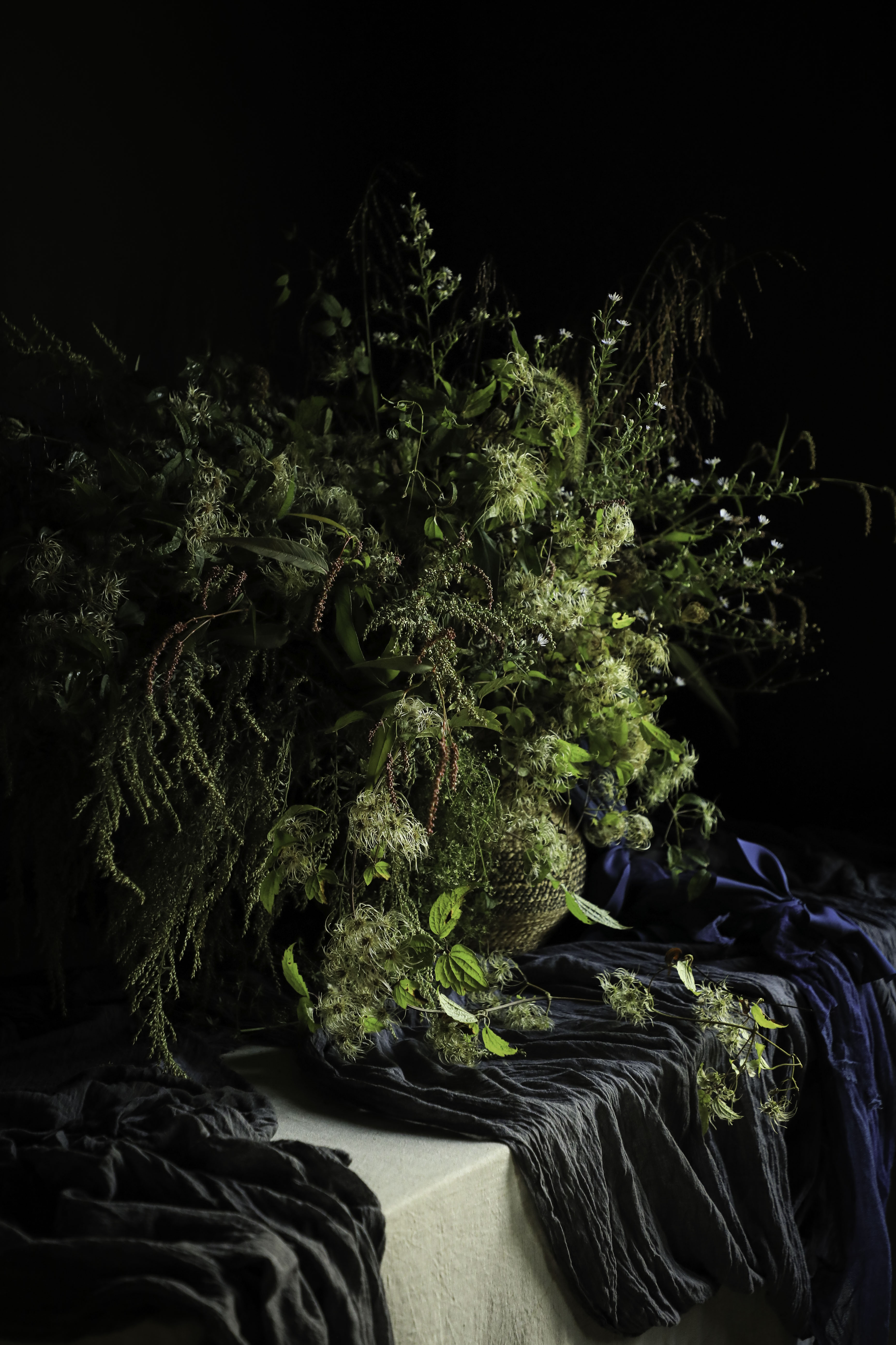 A large, rustic dried flower and botanical arrangement in earthy green and brown tones, styled on a draped dark blue and black fabric surface against a solid black background.