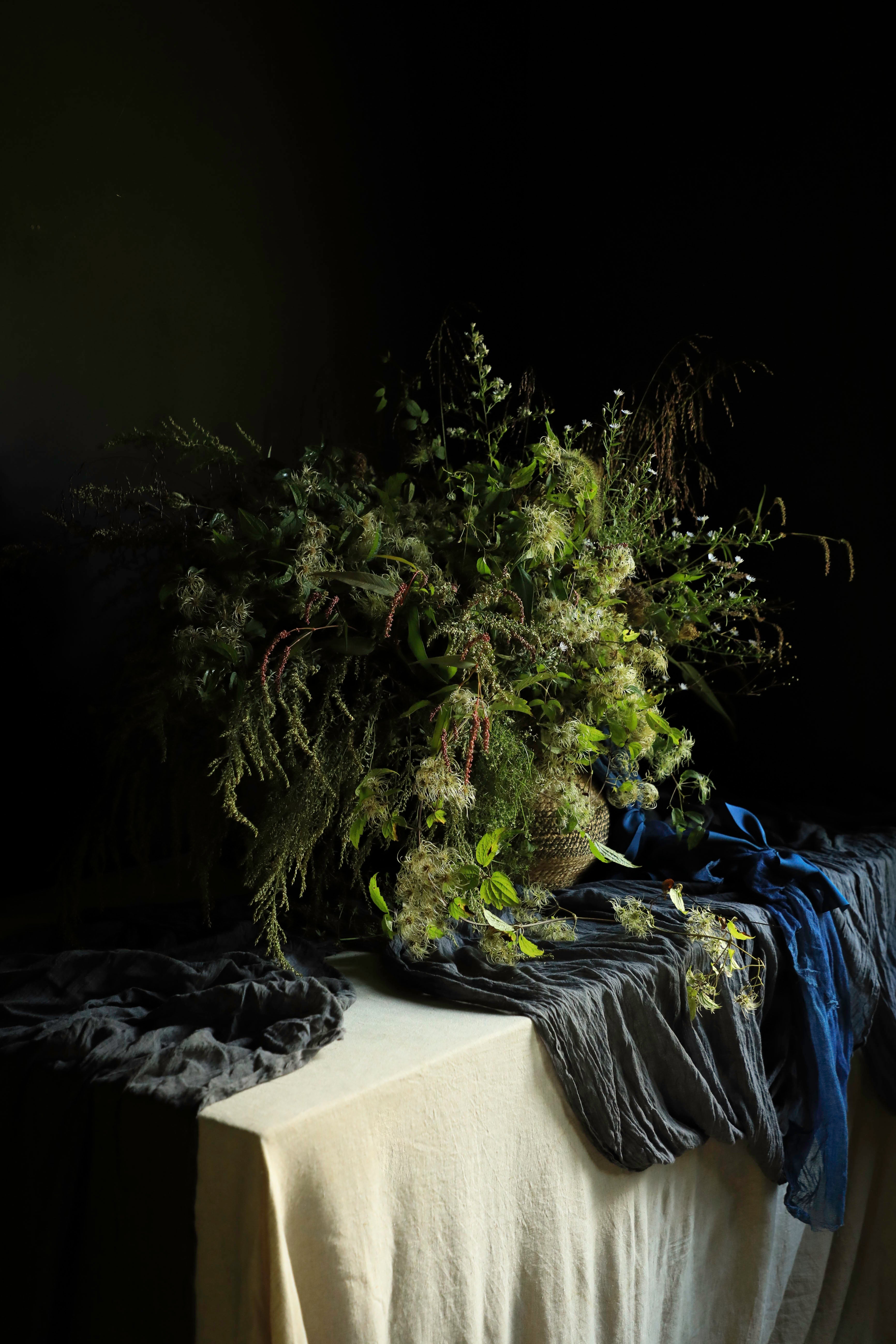 A large, rustic wildflower and foliage arrangement in a woven basket, styled on a draped table with dark gray fabric and a blue ribbon against a dark background.