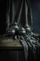 A set of ornate silver metal teapots and cups arranged on a rustic wooden table, draped with dark gray linen fabric against a dark studio background.