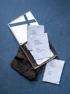 A styled flat-lay of a gold-framed mirror with a white cross design, containing handmade off-white paper cards with printed text, alongside a dark brown textured fabric, all arranged on a textured blue background.