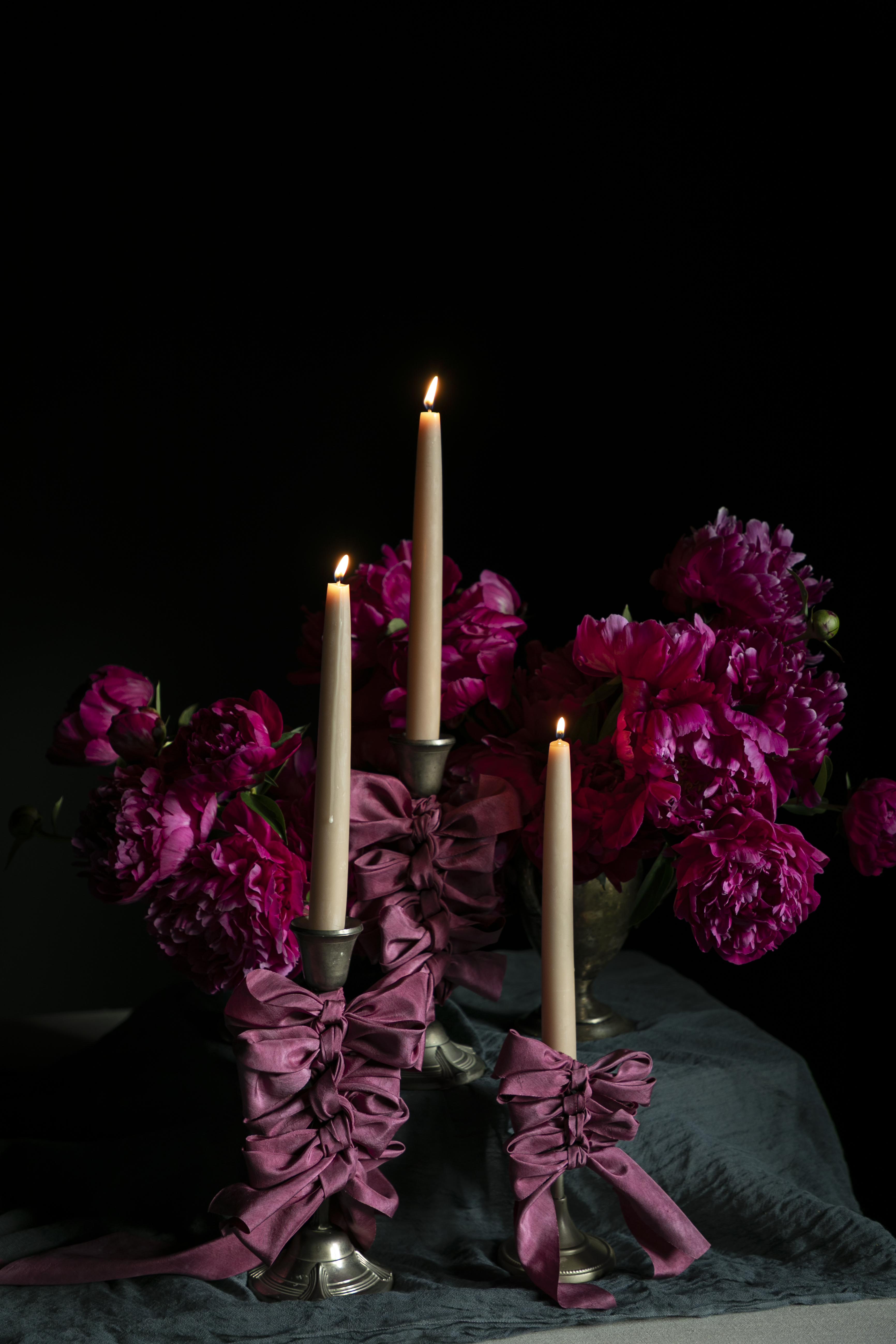 opalite aesthetic. Candle holder with purple ribbons and candles surrounded by pink peony flowers on a dark background. wedding table inspiration