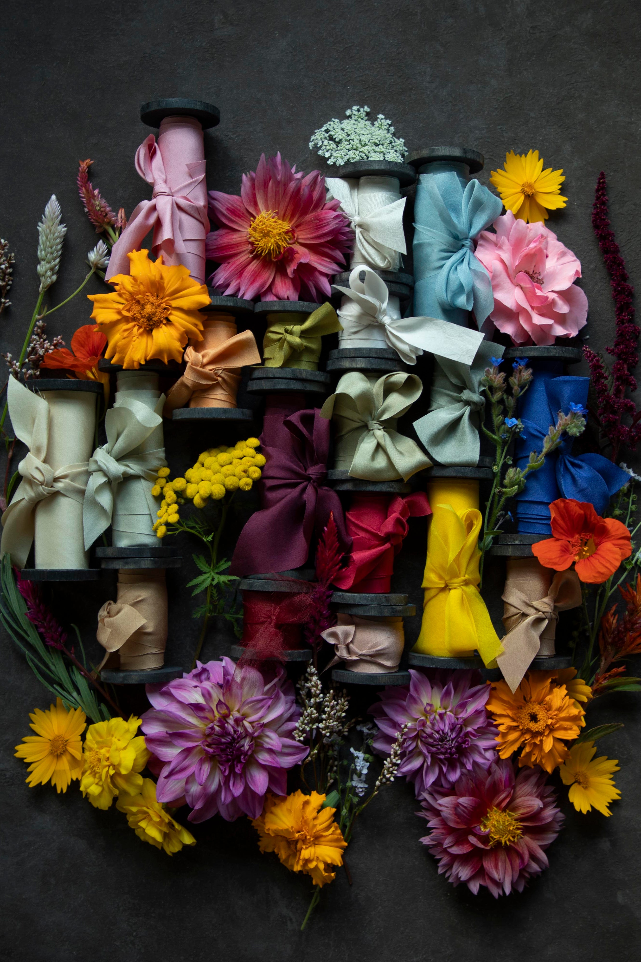 A styled flat-lay of multiple colorful silk ribbon spools wrapped with ribbons and surrounded by vibrant flowers, arranged on a dark textured background.