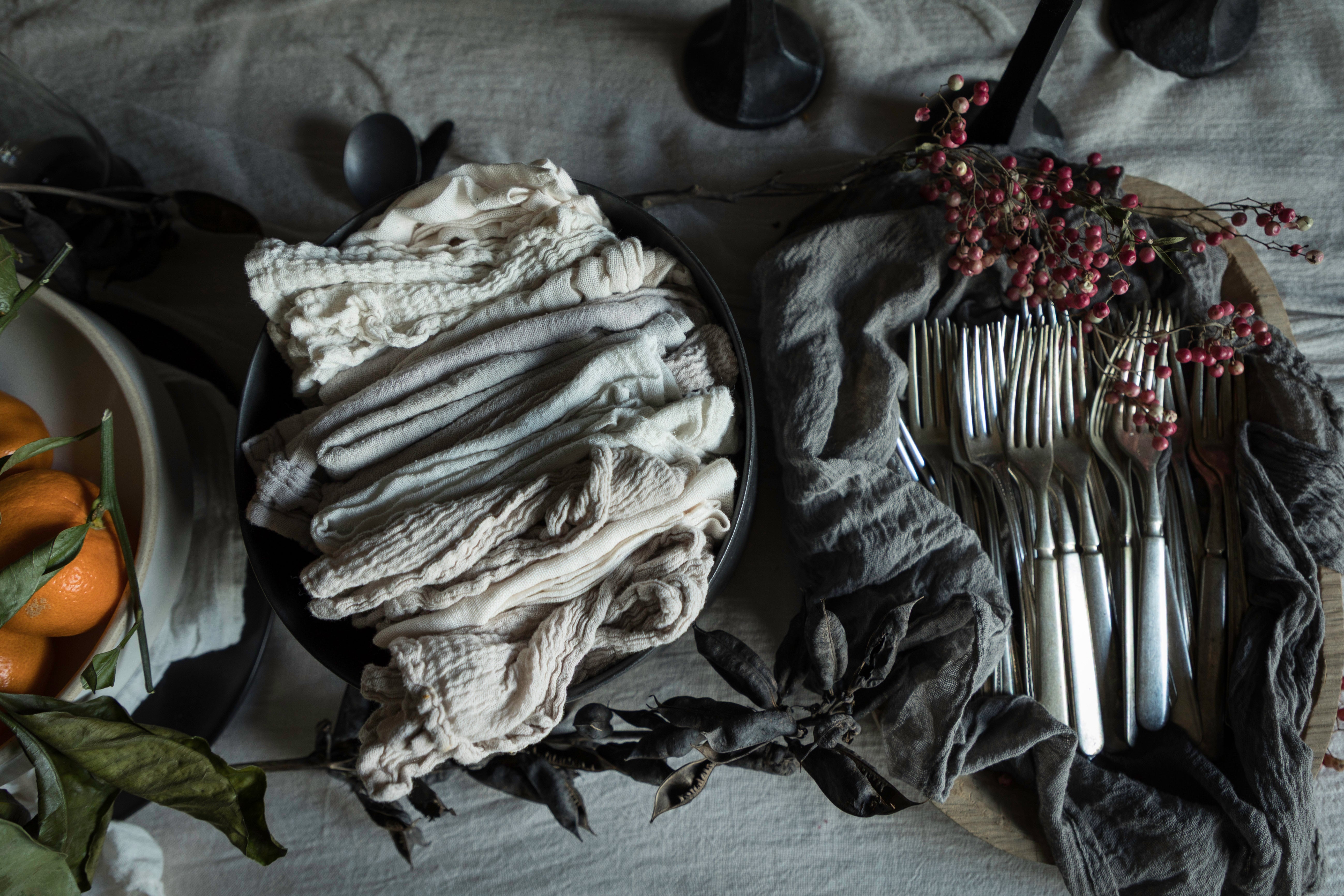 A styled flat-lay of folded fabric napkins or table linens in neutral tones, arranged alongside silver forks on a dark cloth with dried leaves and small red berries on a table covered in a light linen fabric.