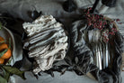 A styled flat-lay of folded fabric napkins or table linens in neutral tones, arranged alongside silver forks on a dark cloth with dried leaves and small red berries on a table covered in a light linen fabric.