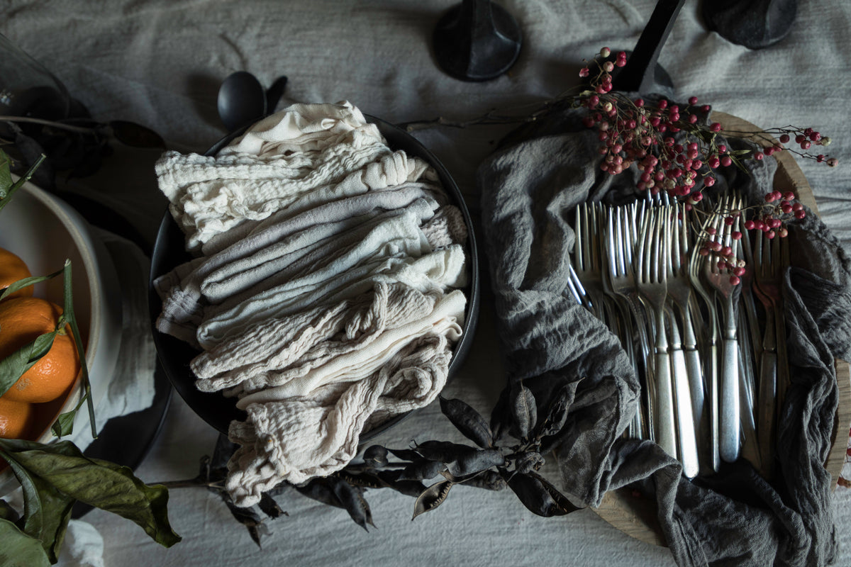 A styled flat-lay of folded fabric napkins or table linens in neutral tones, arranged alongside silver forks on a dark cloth with dried leaves and small red berries on a table covered in a light linen fabric.