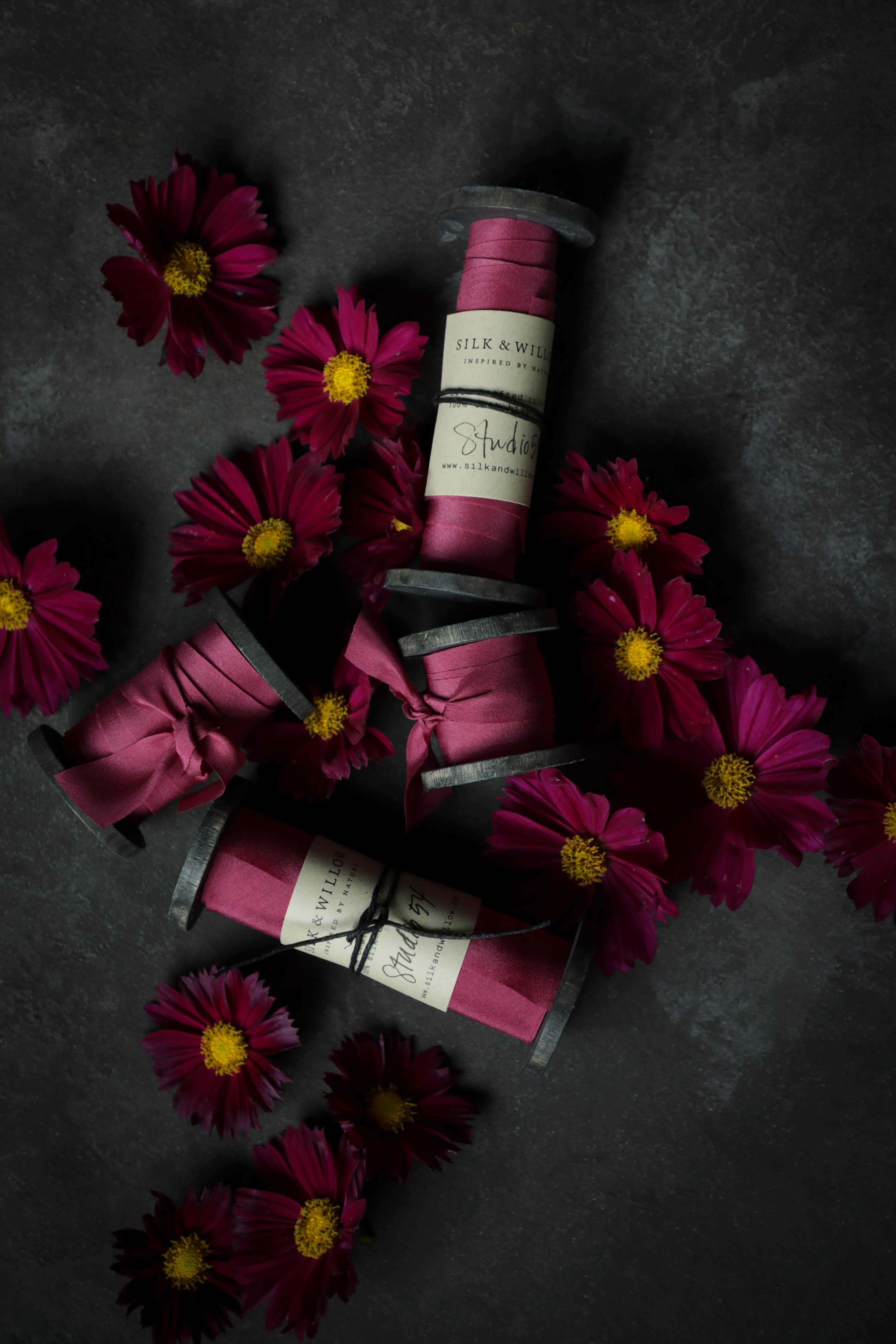 Spools of pink ribbon with labels on a dark surface surrounded by purple flowers.