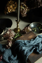 A styled tabletop scene with a crinkled slate-blue fabric draped over a dark wooden board, surrounded by dried flowers, a brass candlestick, and dark ceramic dishes in a moody interior setting.