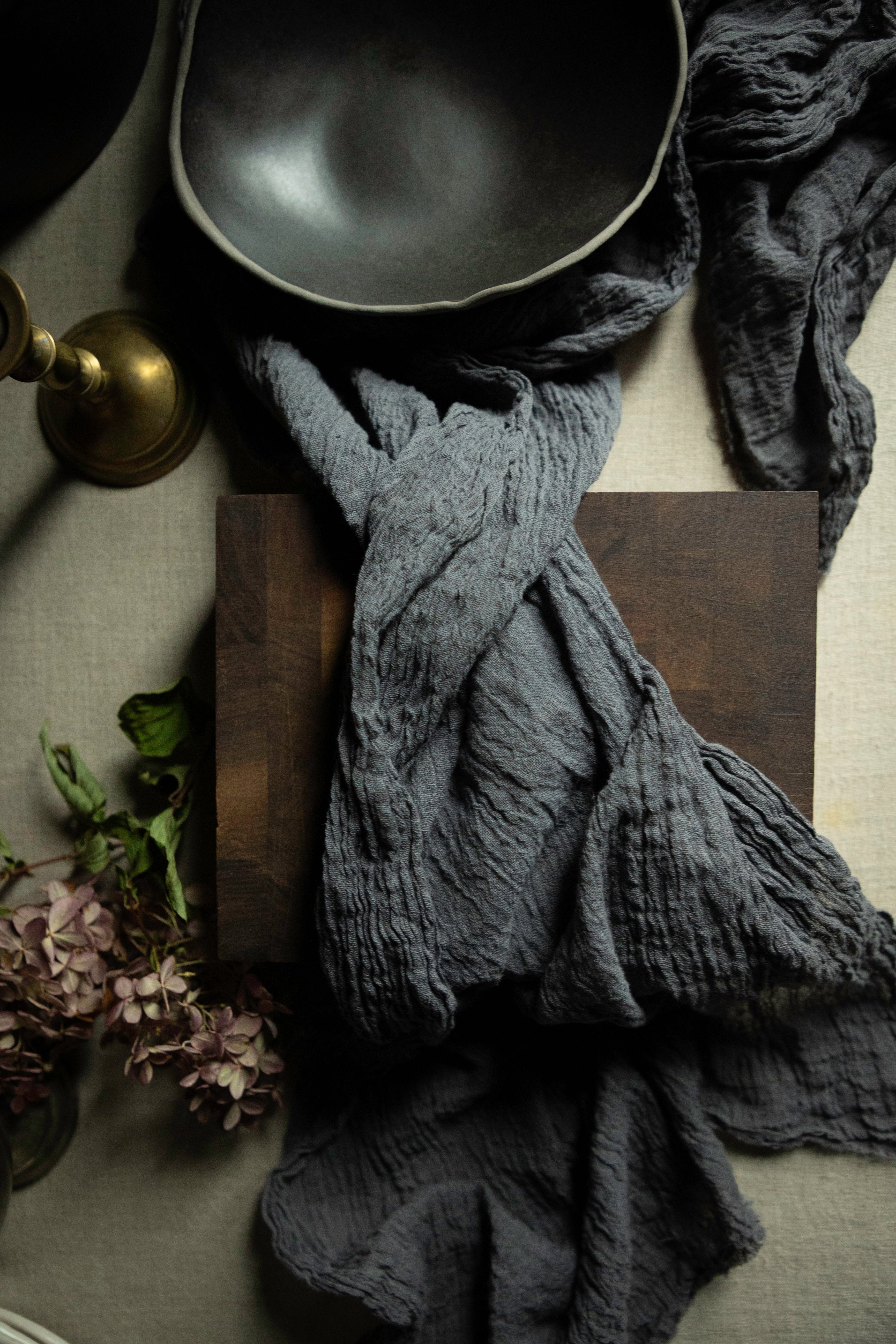 A dark charcoal gray crinkled gauze cotton textile draped over a dark wooden cutting block, styled with a black bowl, a brass candle holder, and dried flowers on a neutral fabric background.