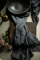 A dark charcoal gray crinkled gauze cotton textile draped over a dark wooden cutting block, styled with a black bowl, a brass candle holder, and dried flowers on a neutral fabric background.