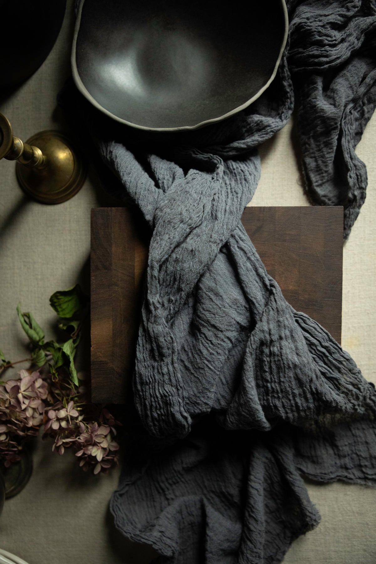 A dark charcoal gray crinkled gauze cotton textile draped over a dark wooden cutting block, styled with a black bowl, a brass candle holder, and dried flowers on a neutral fabric background.